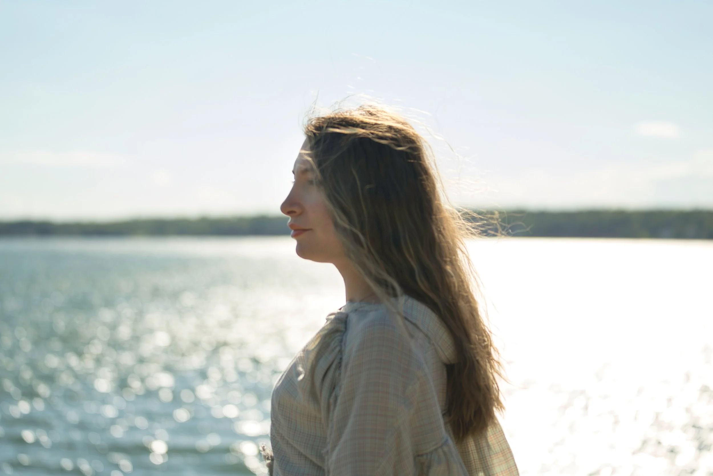 A woman with long hair standing by a body of water during sunset with her eyes closed.