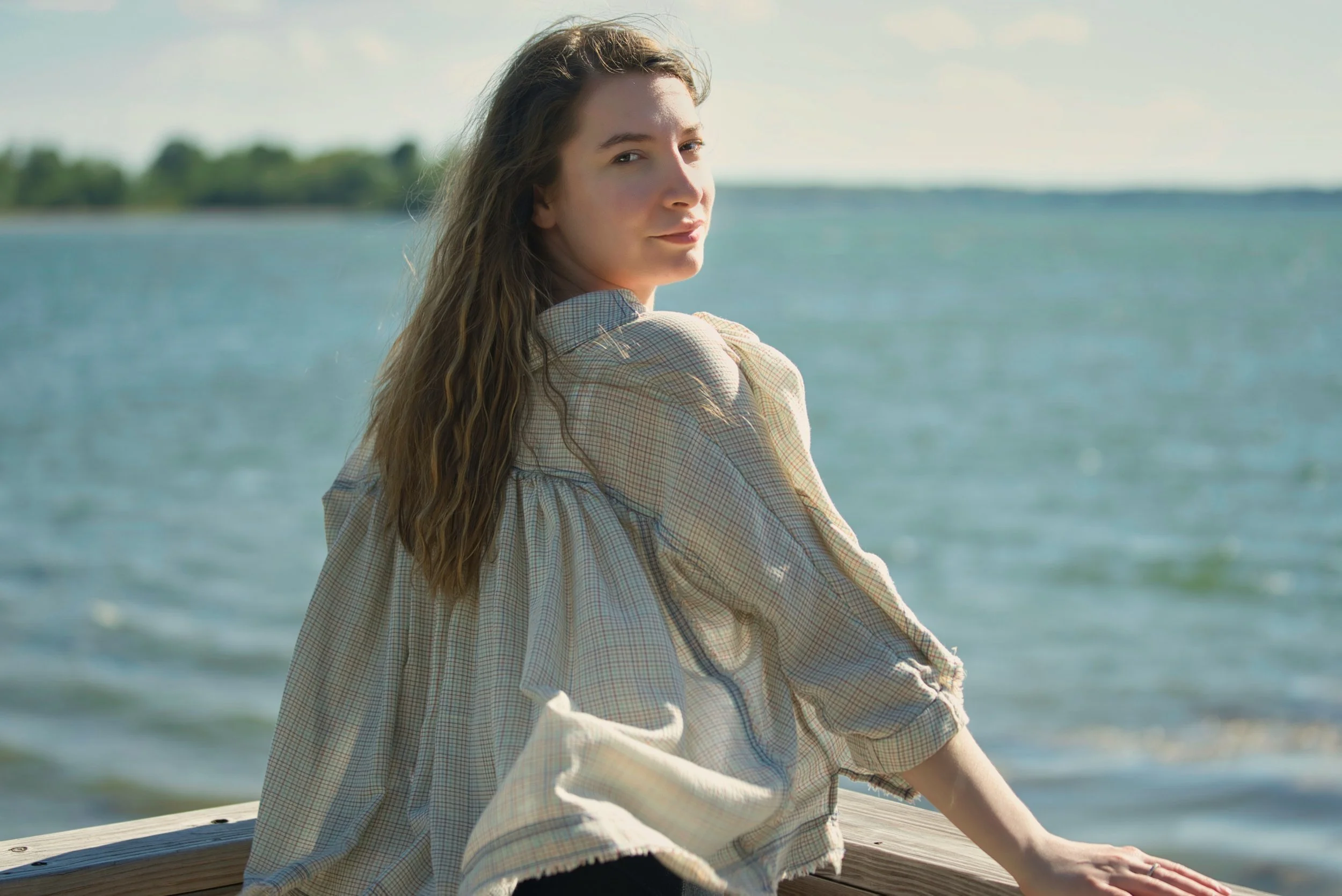 A young woman with long wavy hair wearing a plaid shirt, sitting on a wooden dock near a body of water, looking at the camera with a slight smile, during daytime.