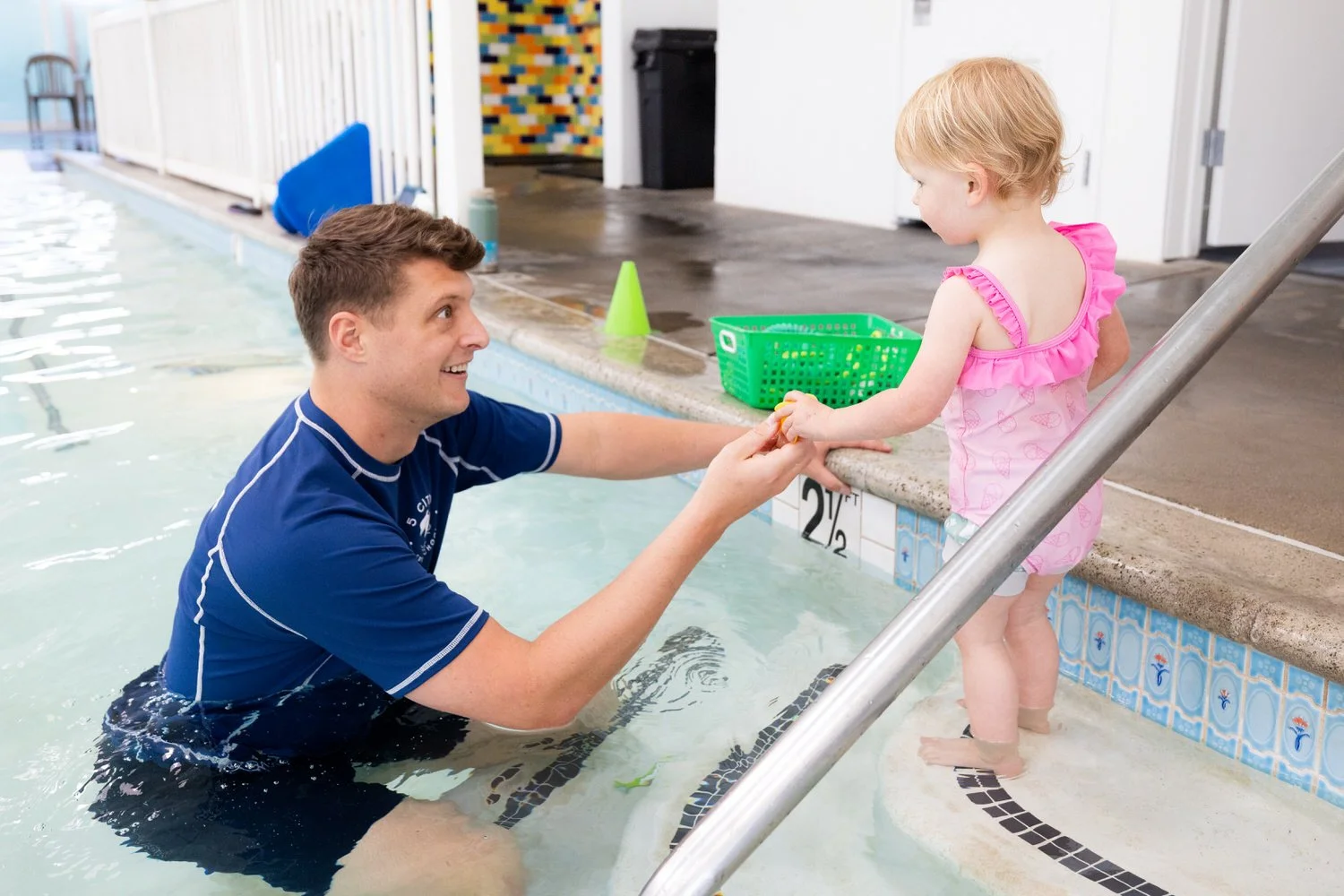 A young man in a blue swim shirt is helping a small girl in a pink bathing suit out of a swimming pool. The girl is standing in shallow water and holding a yellow toy, while the young man is holding her hands and smiling at her. The pool area includes a green basket, a lane line, and a wall with colorful tiles in the background.