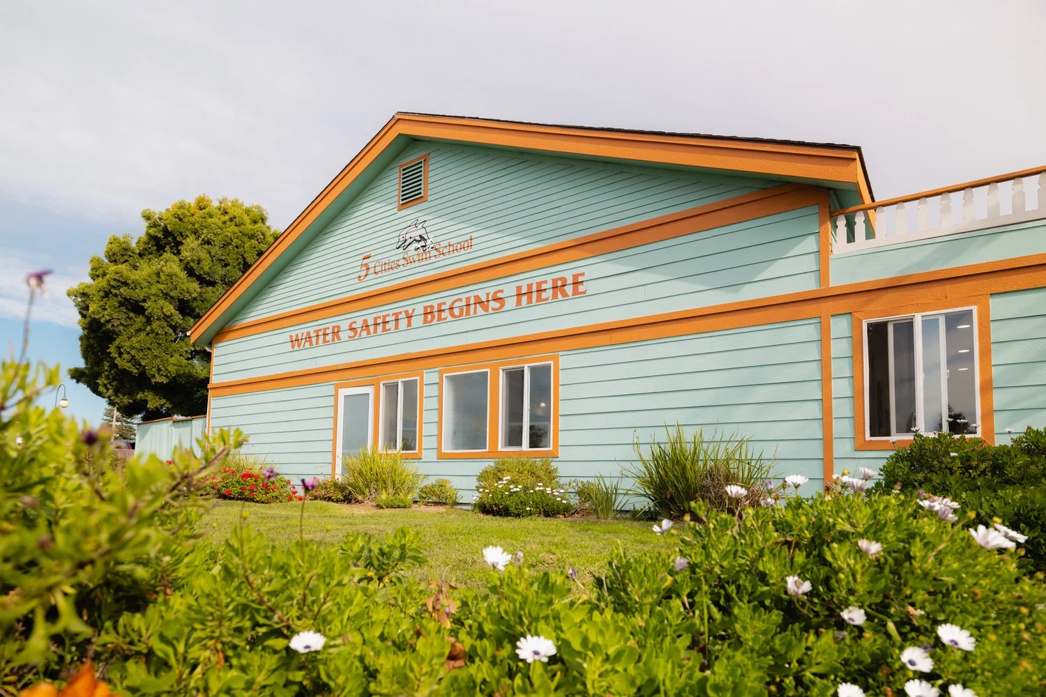 A colorful building with teal walls and orange trim, labeled '5 Cites Swim School,' with a sign that reads 'Water Safety Begins Here,' surrounded by green plants and flowers.