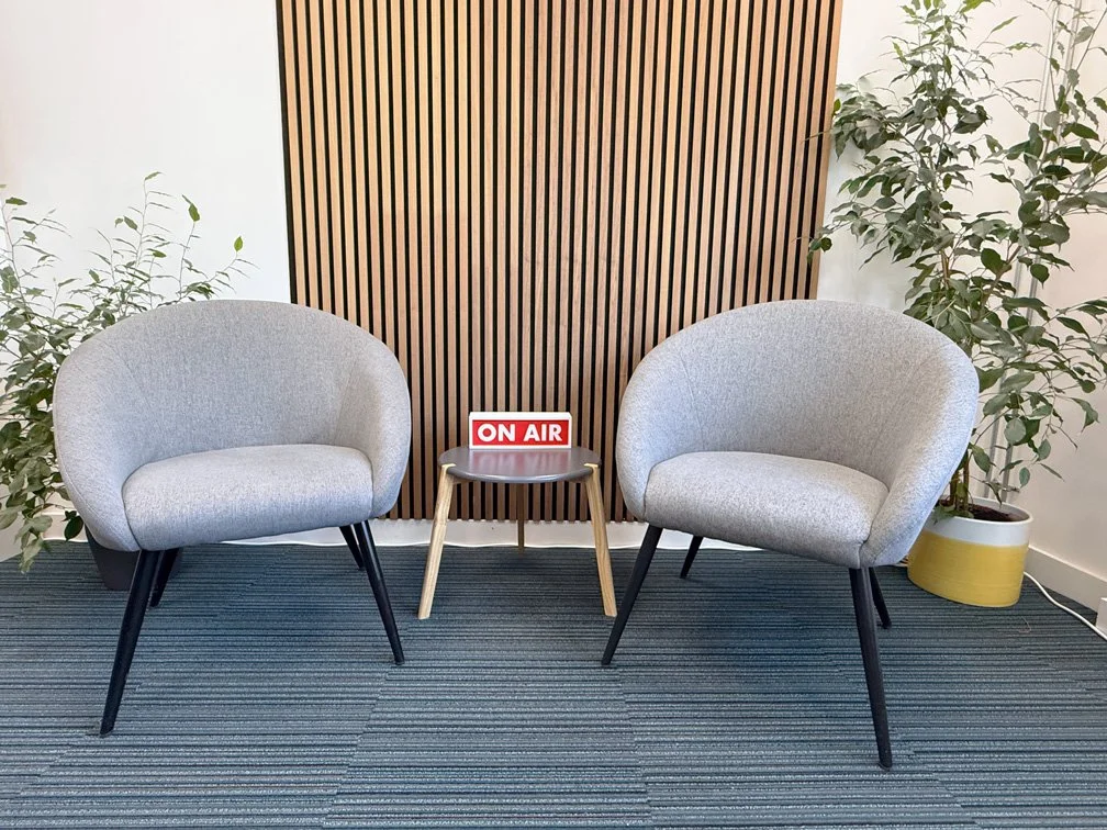 Two grey upholstered chairs with black legs and a small wooden table in between, featuring a red and white 'ON AIR' sign, situated in a modern room with wooden slat wall, potted plants on either side, and striped carpet flooring.
