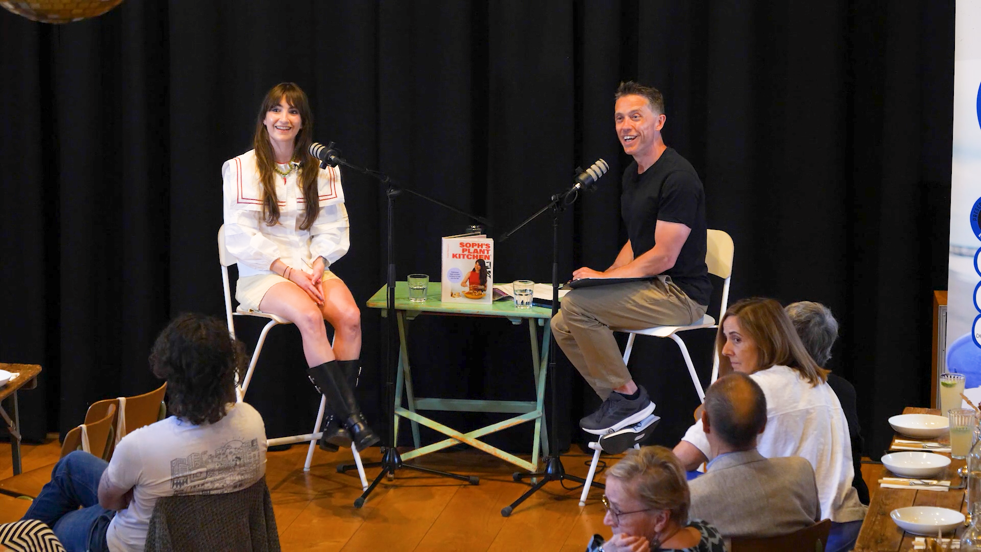 A woman and a man sit on stage engaged in a conversation during a book talk. The woman is dressed in a white outfit with long brown hair, and the man is in a black shirt and tan pants. There are microphones, glasses of water, and a book titled 'Sophi's Plant Kitchen' on a small table between them. The audience sits in front, listening to the discussion.