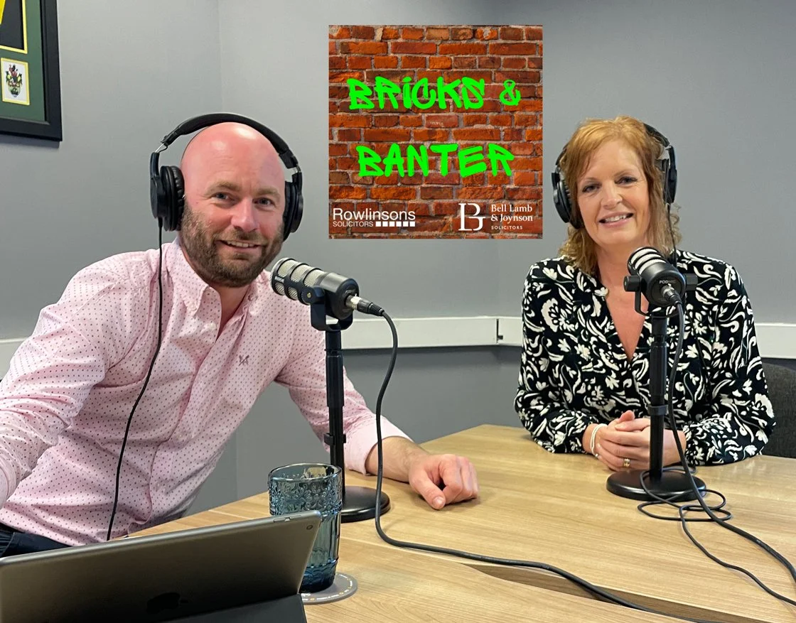 Two people, a man and a woman, sitting at a table with microphones, headphones, and a laptop in a podcast studio. Behind them is a sign that says 'Bricks & Banter' with a brick wall background and logos of Rowlinsons Solicitors and Bell Lamb & Joynson Solicitors.