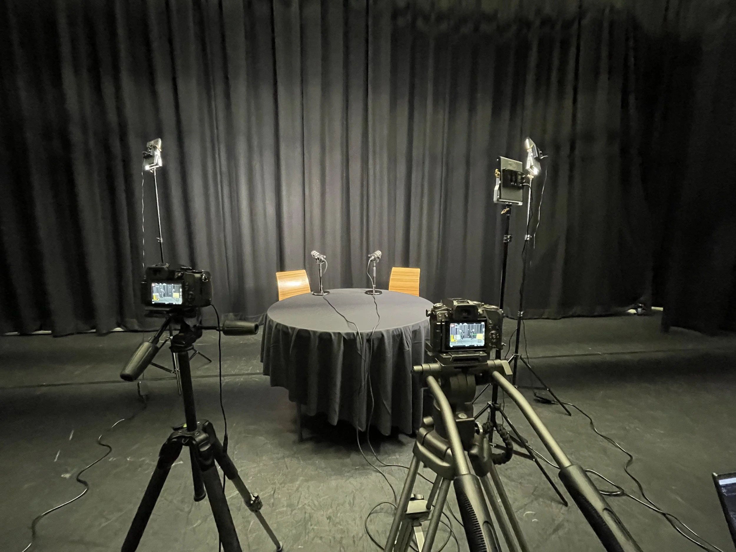 Empty stage with a black curtain backdrop, a table with two microphones and two wooden chairs, and cameras on tripods pointing toward the stage, set up for a presentation or panel discussion.