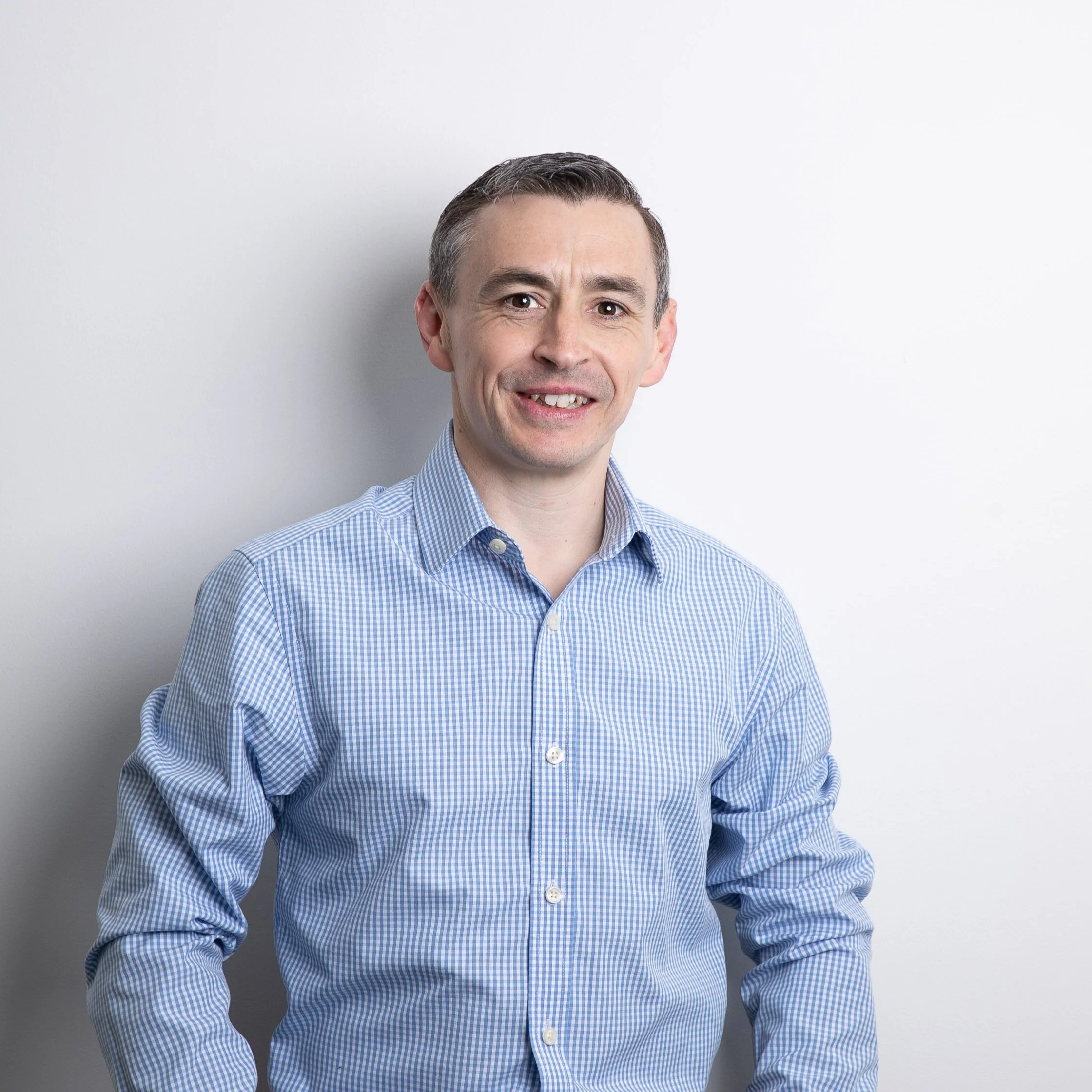 A man with short dark hair smiling, wearing a light blue checked shirt, standing against a plain white wall.