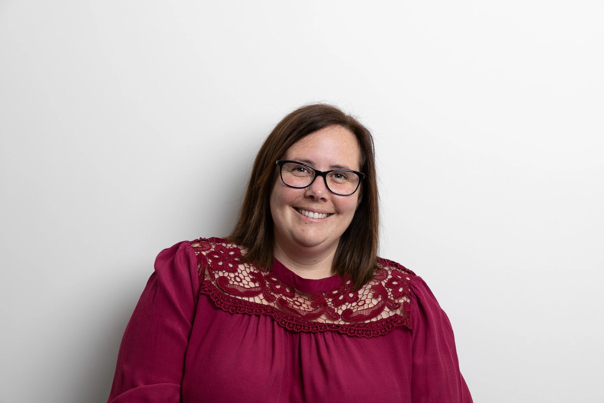 A woman with shoulder-length brown hair, wearing glasses and a maroon blouse with lace details, smiling while standing against a plain white wall.