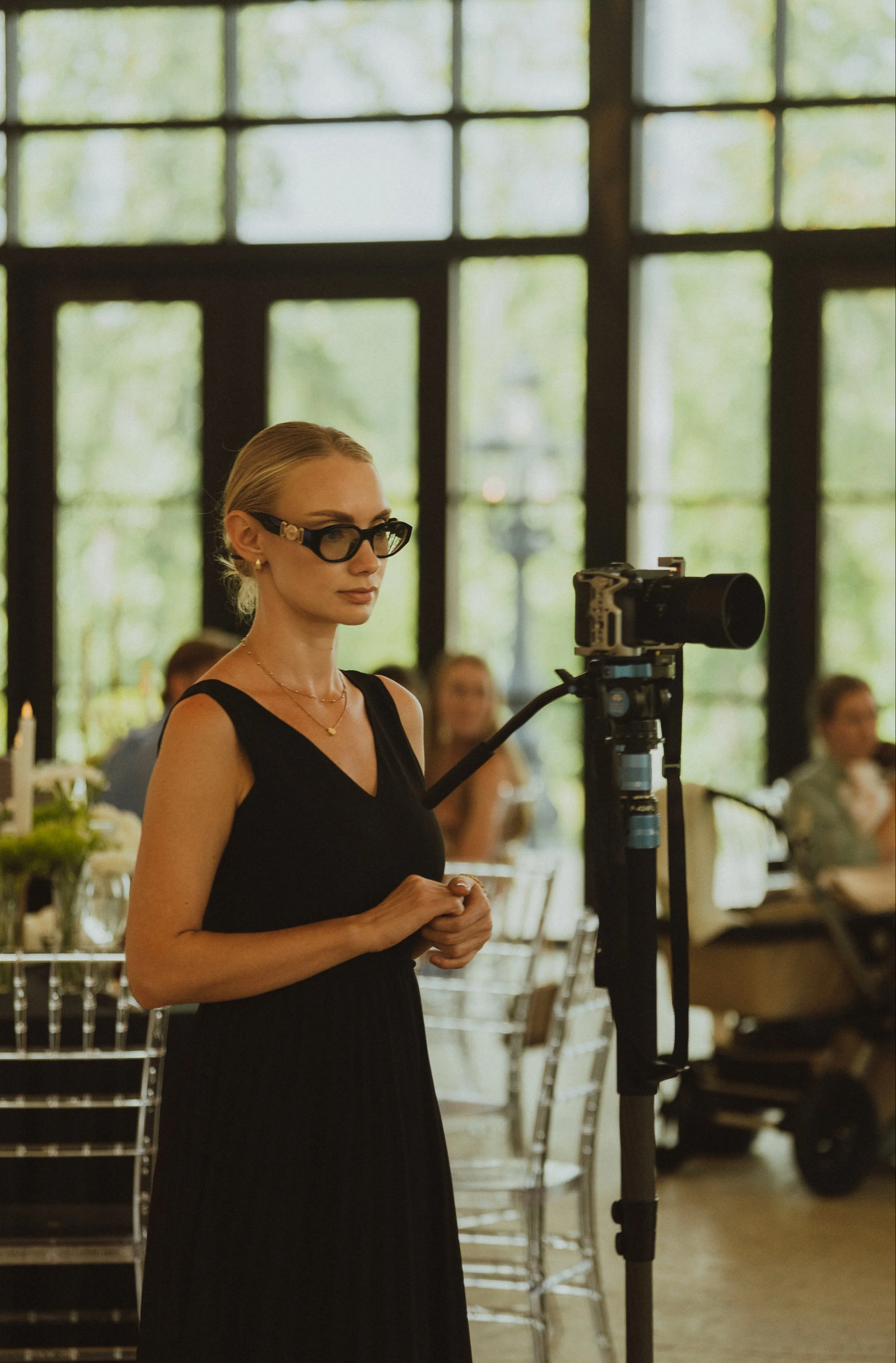 Wedding videographer Valentyna Tiurkin. A woman with blonde hair, wearing glasses and a black sleeveless dress, stands in front of a camera on a tripod, inside a bright room with large windows and tables set for a gathering.