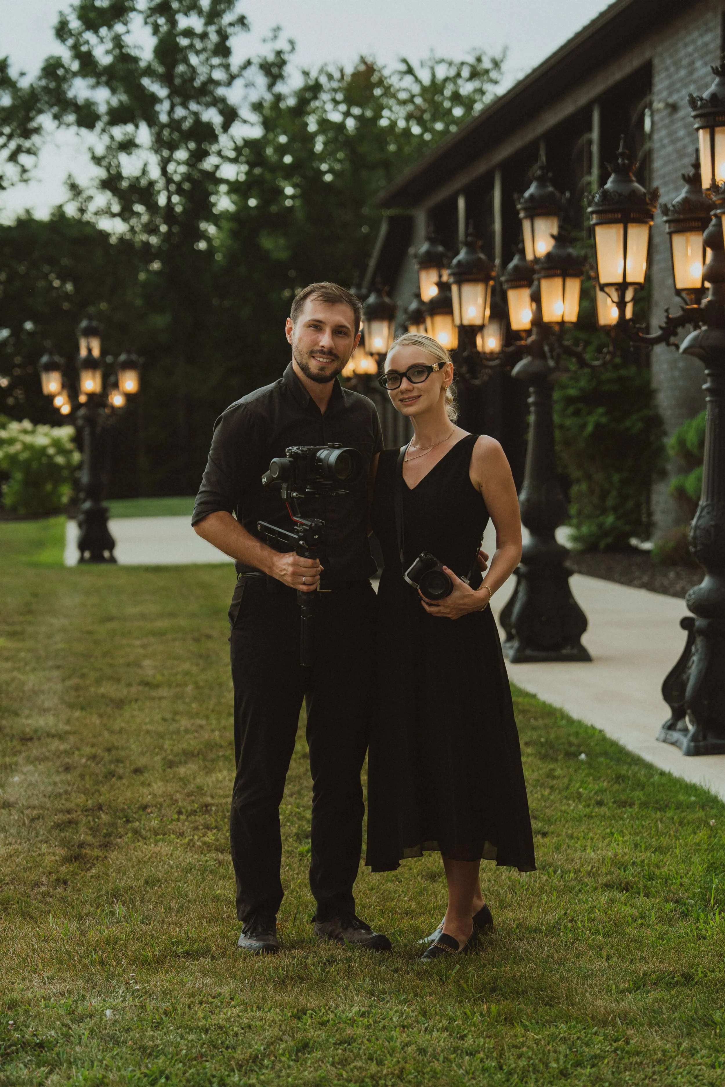Wedding Videographers Oleh and Valentyna Tiurkin standing outdoors at dusk, holding cameras, with large ornate street lamps behind them, a house, and trees in the background.