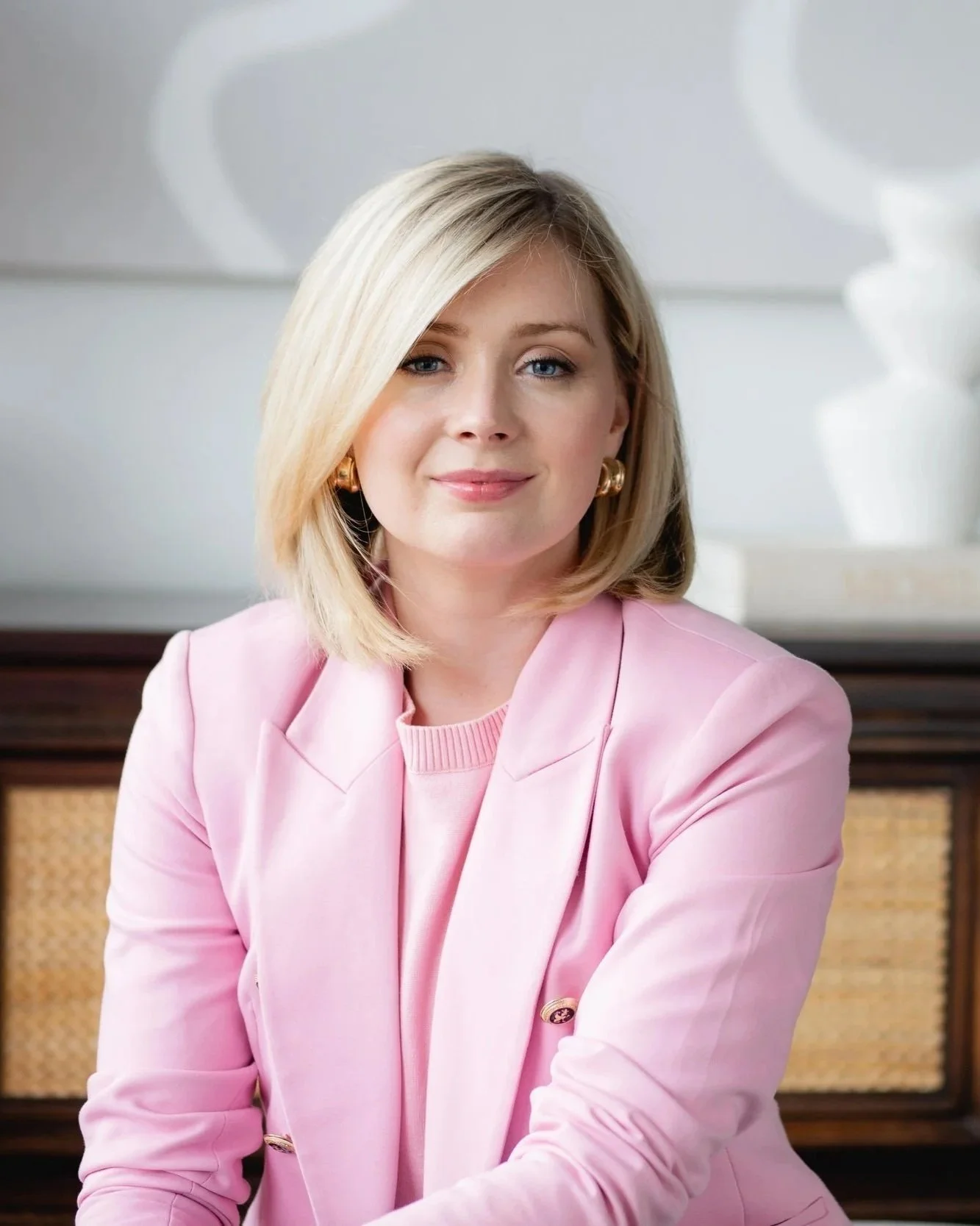 Portrait of a woman with shoulder-length blonde hair, wearing a pink blazer and gold earrings, sitting in front of a white wall with abstract artwork.