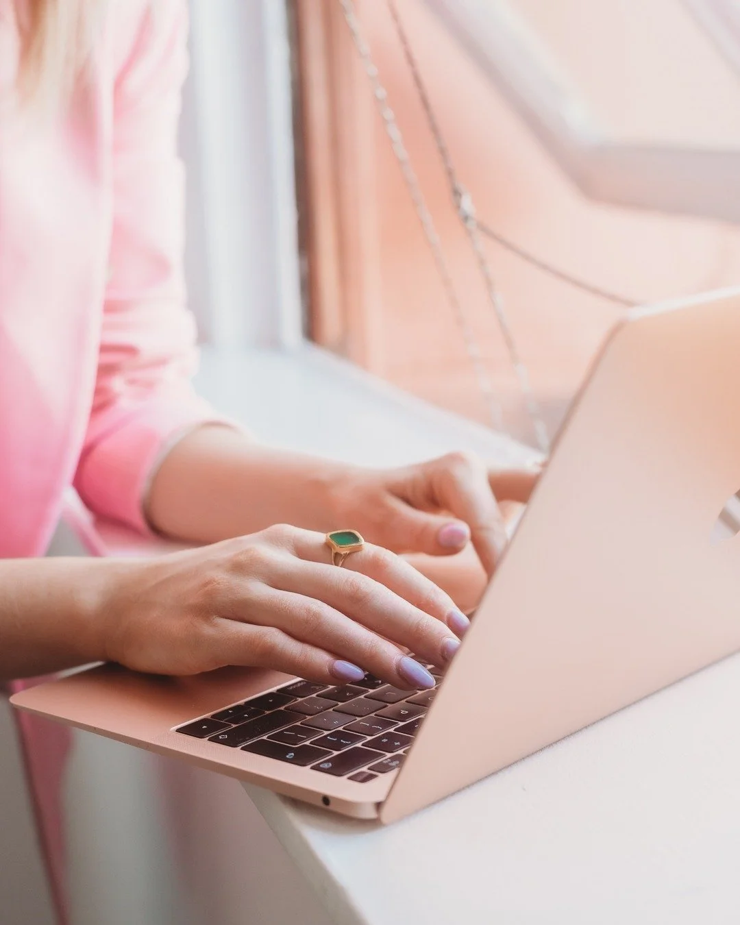 Person wearing a pink sweater typing on a pink laptop on a white table, near a window with pink curtains.