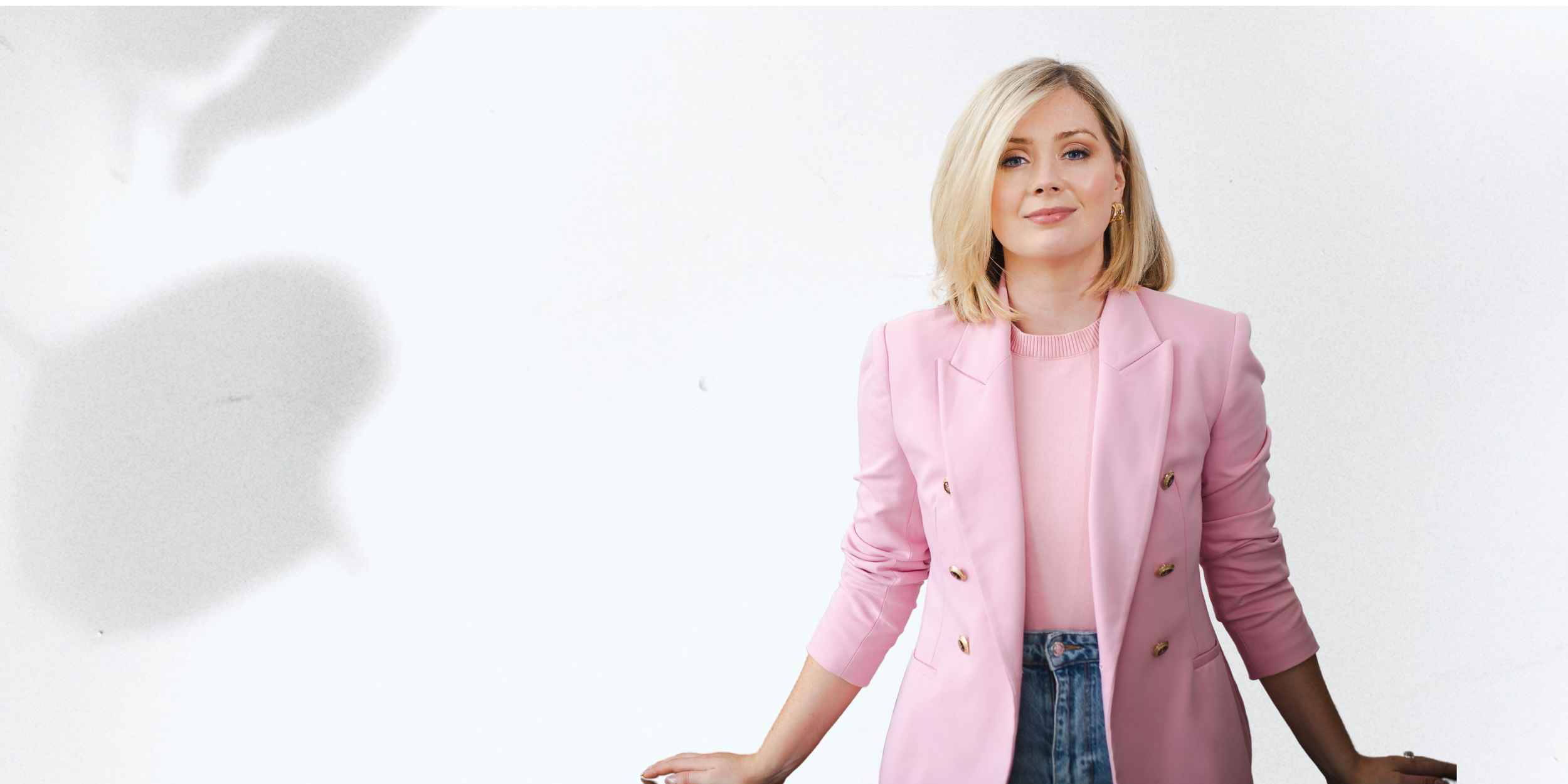 A woman with blonde hair in a pink blazer, pink sweater, and blue jeans standing against a white background.
