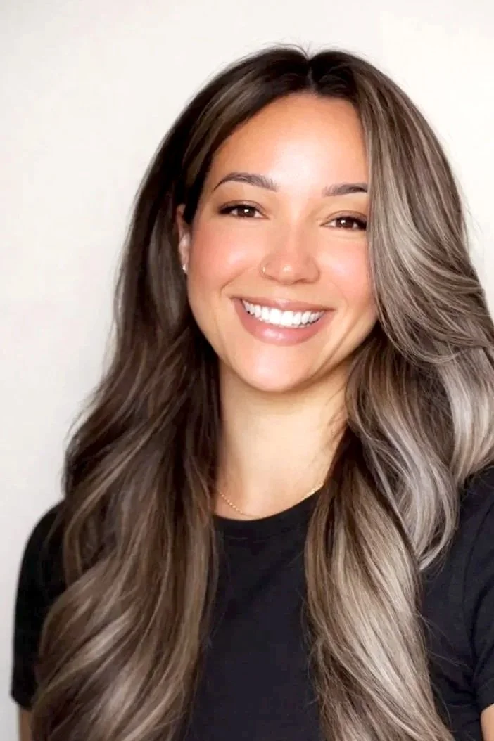 A young woman with long, wavy brown hair and light skin smiling at the camera, wearing a black shirt and a small nose piercing.