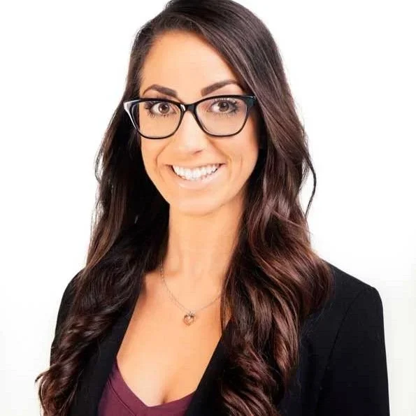 Smiling woman with long wavy brown hair, wearing glasses, a black blazer, and a necklace with a small pendant.