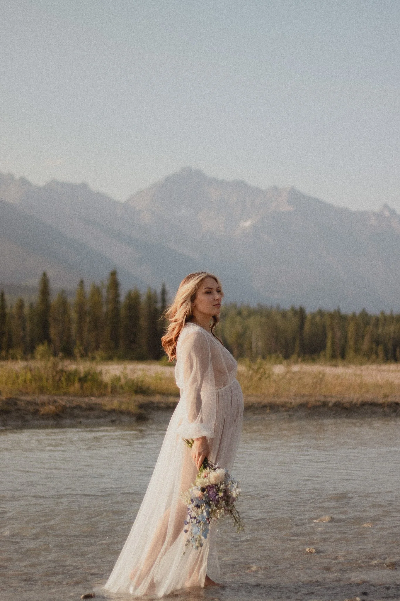 Maternity session. A woman stands in a shallow creek, with the majestic Rocky Mountains behind her. She's holding a stunning bouquet of flowers from Golden floral co.