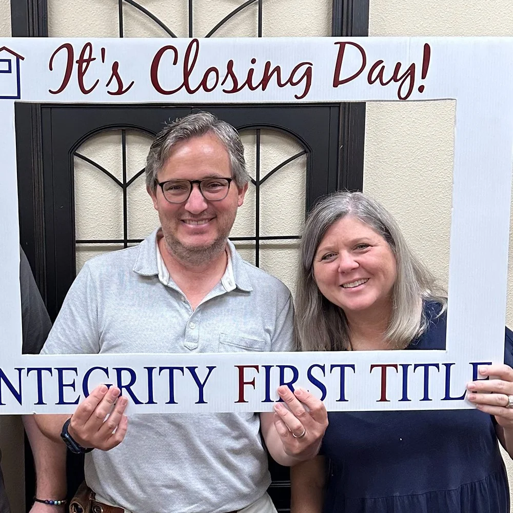 A smiling man and woman holding a sign that says, 'It's Closing Day! INTEGRITY FIRST TITLE,' standing in front of a decorative black door frame with a sign above them.
