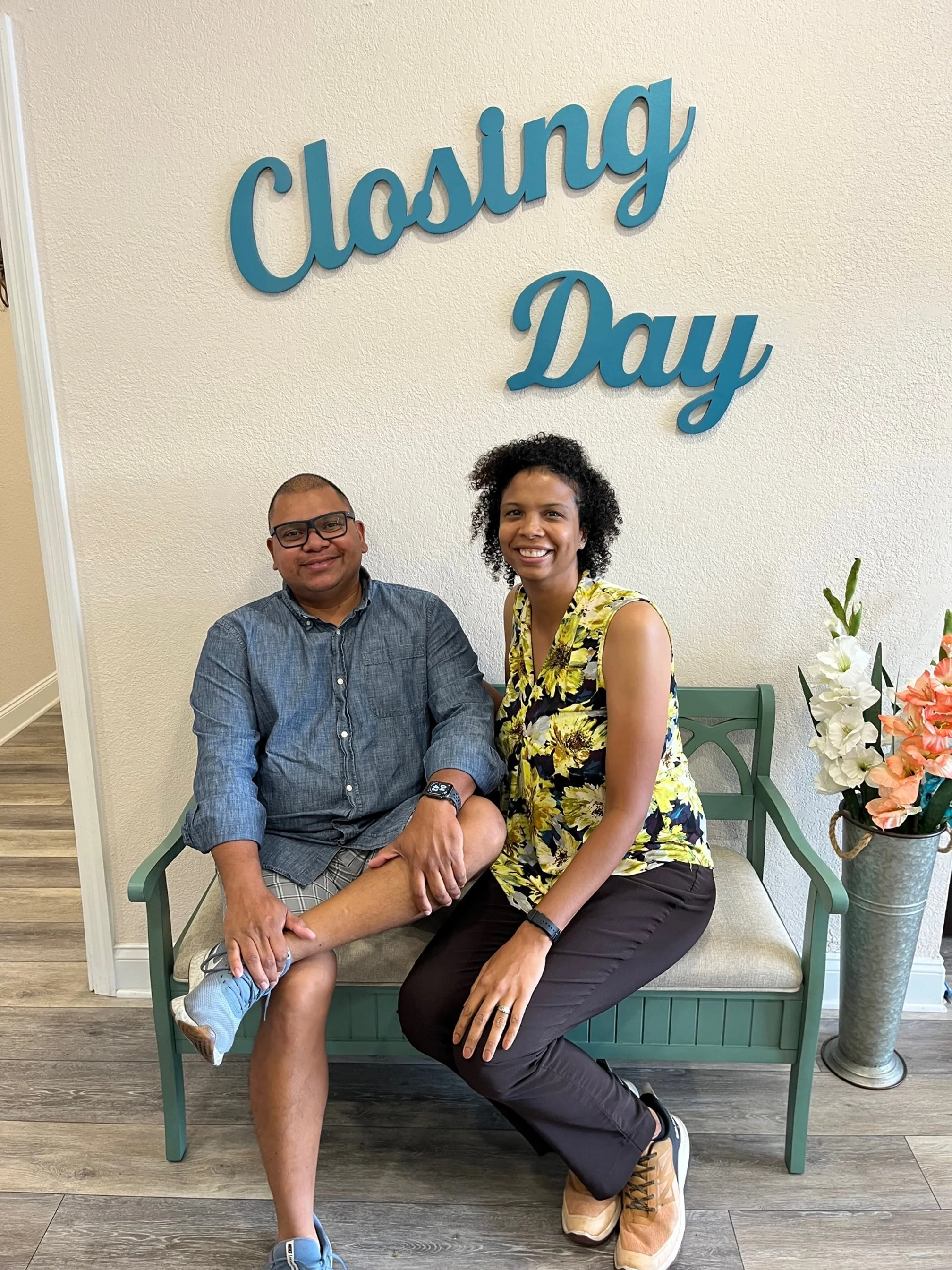 Two people sitting on a bench under a sign that says 'Closing Day.' One person with glasses, wearing a blue shirt, and the other in a floral top and black pants. There are flowers in a vase on the right.