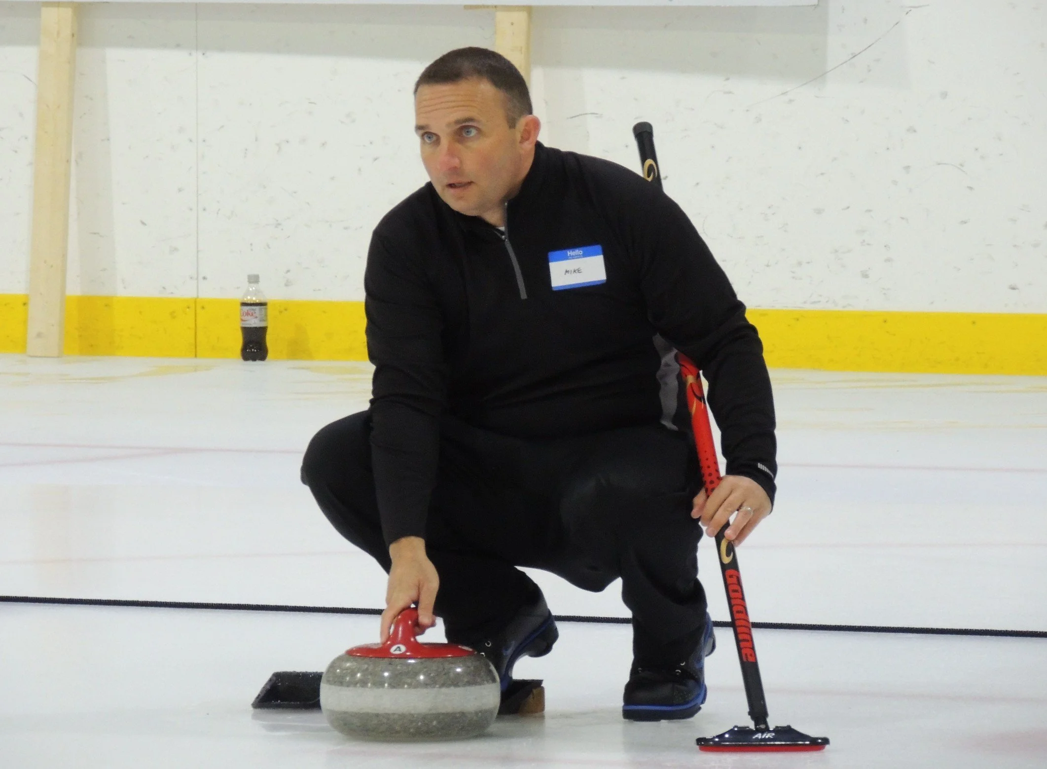 A man kneeling on ice at a curling rink, preparing to slide a curling stone forward with a broom in his right hand, wearing black sportswear with a name tag labeled "Mike."
