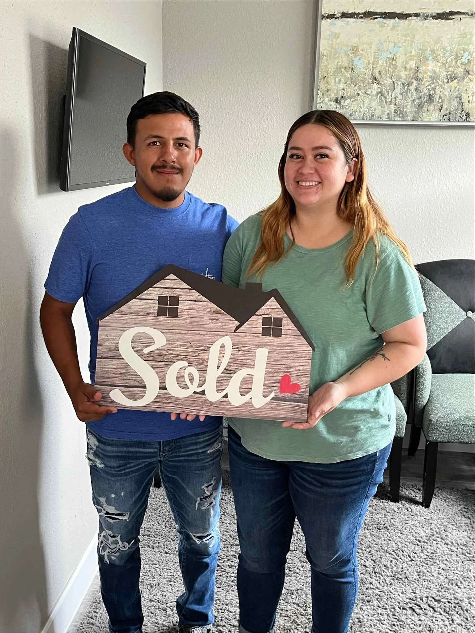 A couple standing inside a living room holding a wooden house-shaped sign that reads "Sold" with a small red heart. The man wears a blue shirt and ripped jeans, and the woman wears a green shirt and jeans. They are smiling.