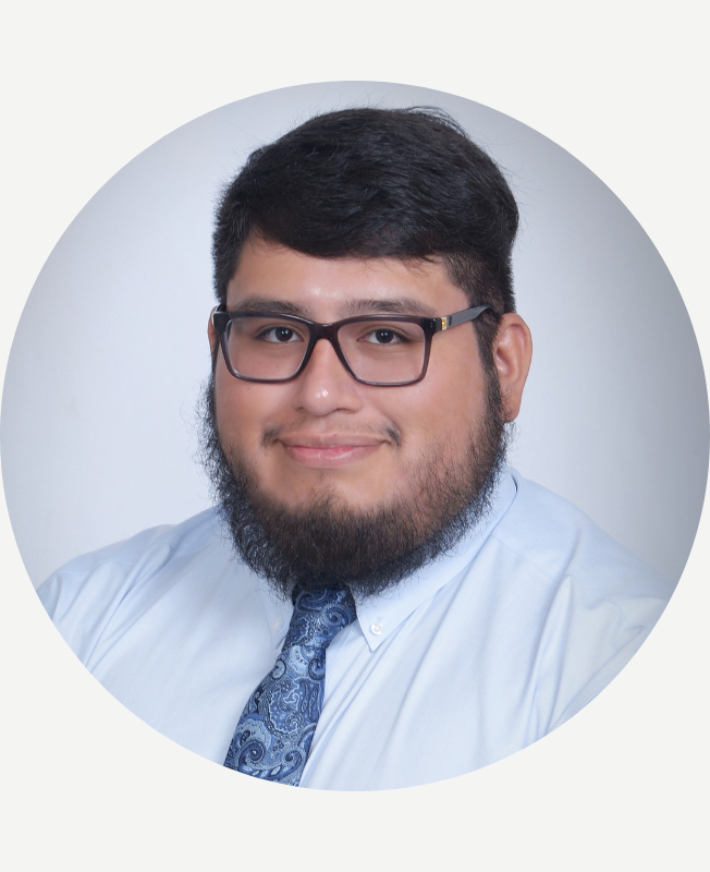 Headshot of a young man with glasses, black hair, beard, wearing a light blue shirt and patterned blue tie, smiling against a white background.