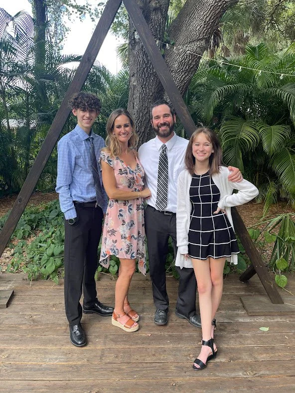 A group of four people, two women and two men, standing outdoors on a wooden platform with lush green plants and trees in the background. They are smiling and posing for a photo.