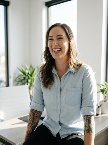 A woman with tattoos on her arms, wearing a light blue button-up shirt, laughing in a bright office with large windows and green plants in the background.