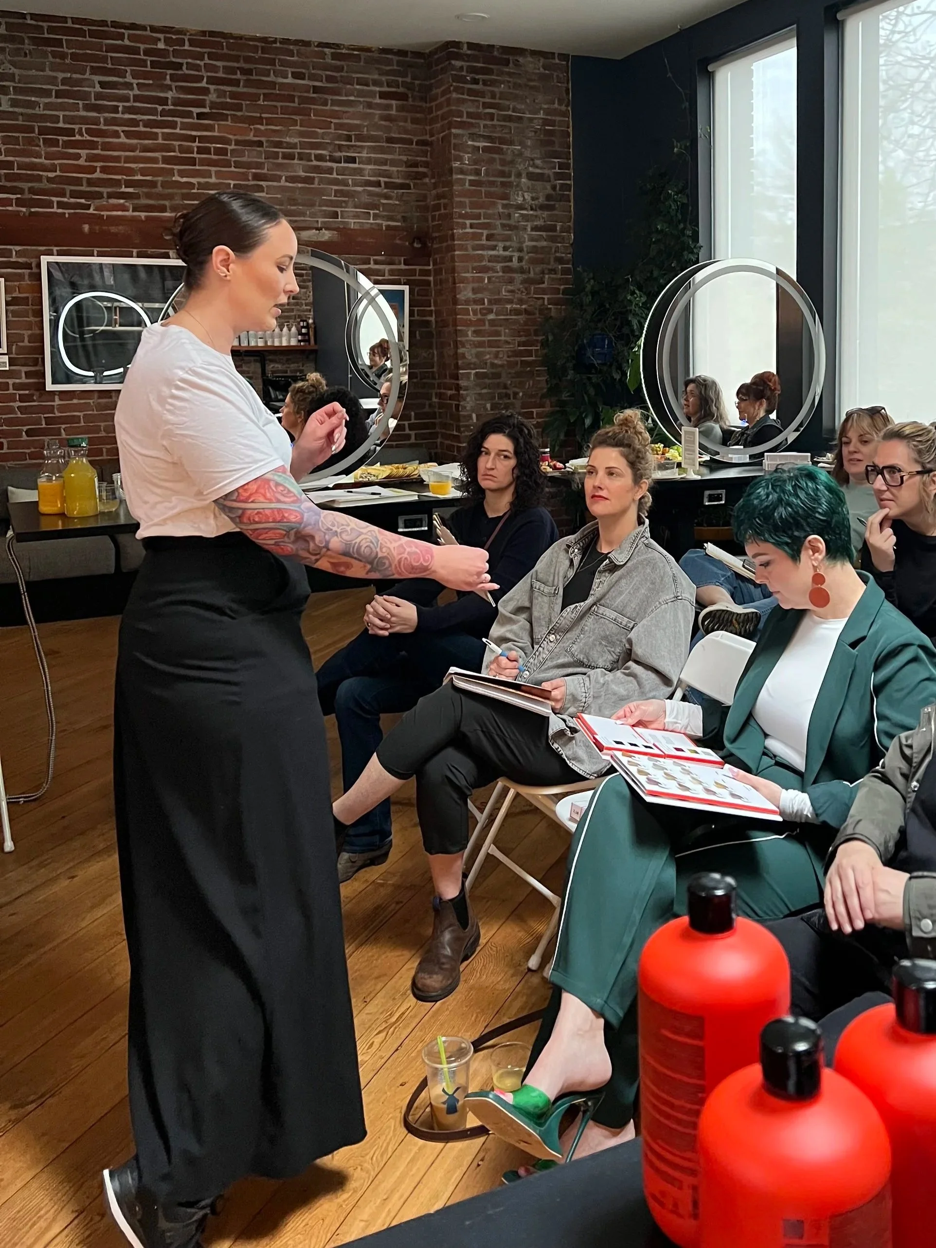 A woman with dark hair and tattoos on her arms is standing in a salon with a group of women seated, some taking notes or listening attentively. The salon has exposed brick walls, large windows with white blinds, and bottles of hair product on a nearby table.