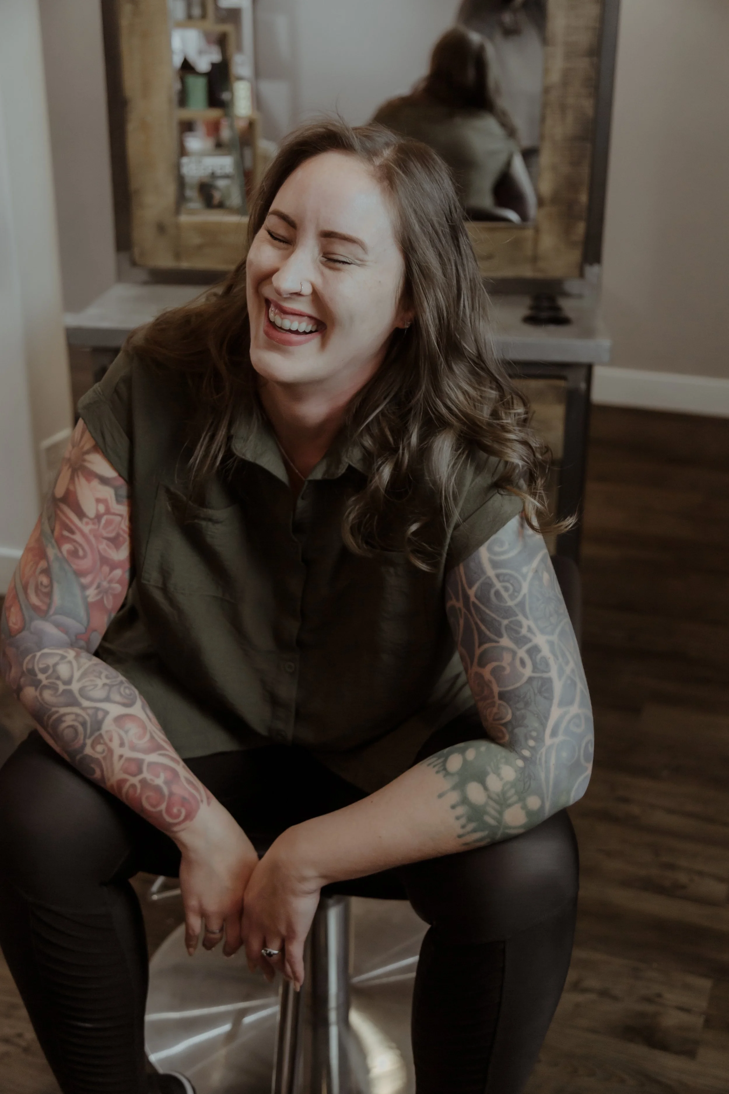 A woman with long brown hair and tattoos on her arms, smiling and laughing, sitting on a stool in a room with wooden floors and a mirror in front of her.