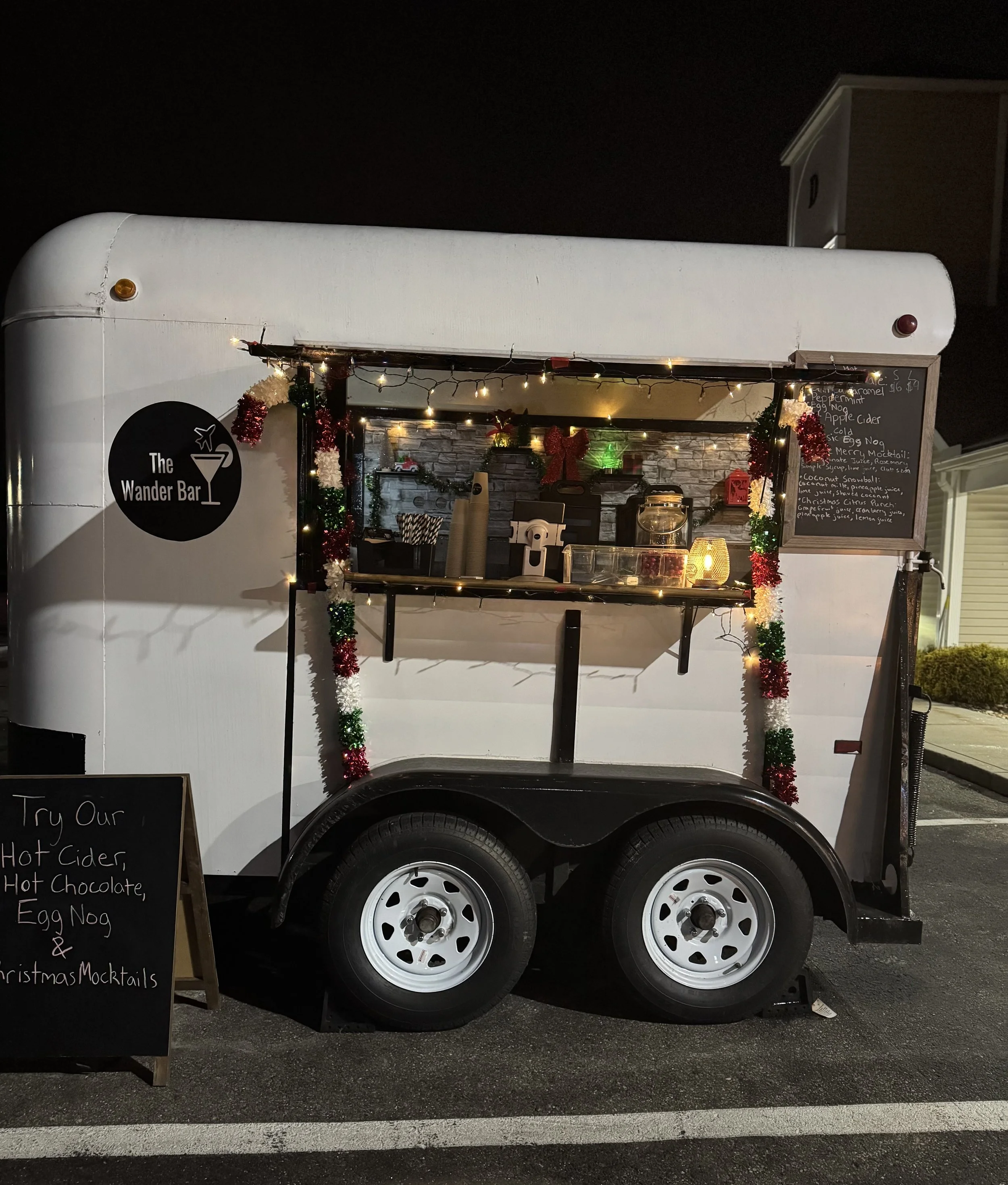 Vintage horse trailer bar is decorated with Christmas lights and candy canes with a sign out front advertising mocktails and beverages in Londonderry New Hampshire