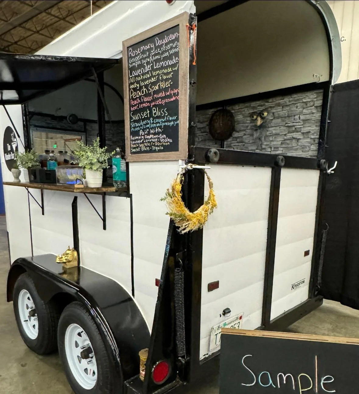 The Wander Bar vintage horse bar trailer is decorated with florals as they advertise sample mocktails from their custom mocktail menu at a wedding show event in Wilmington MA