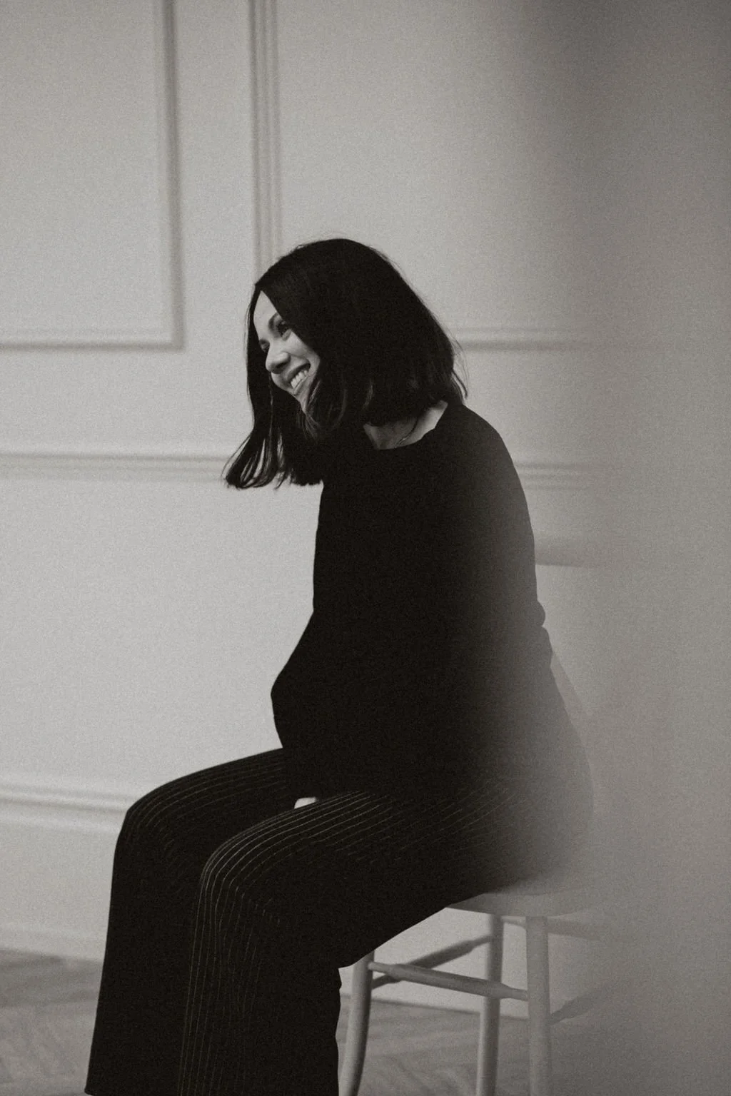 A woman with shoulder-length hair smiling while sitting on a chair in a room with plain walls, captured in black and white.