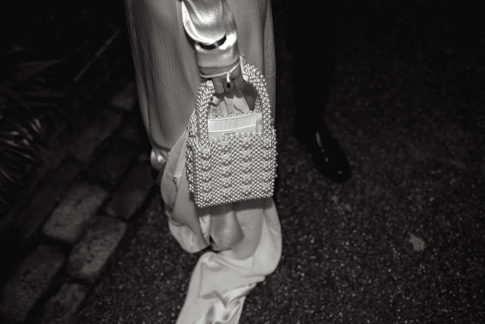 A person holding a pearl handbag, dressed in light-colored clothing, standing on a dark asphalt surface near a brick wall.