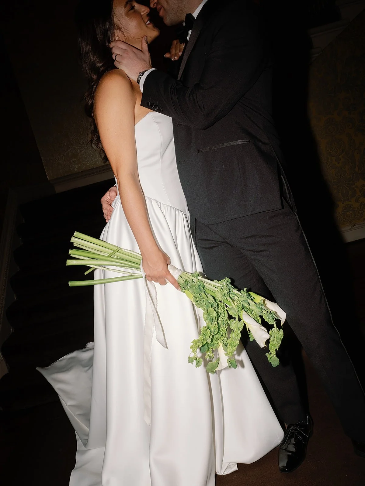 A bride in a white wedding gown holding a bouquet of green flowers, with a groom in a black tuxedo, sharing an intimate moment at their wedding.