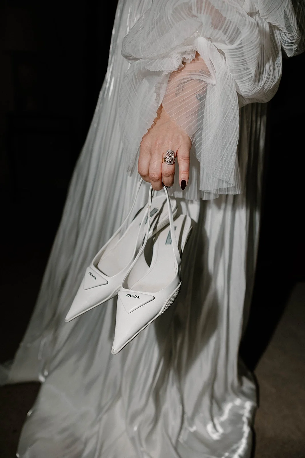 Close-up of a woman's hand holding a pair of white Prada high heels, wearing a large silver ring, with sheerness and pleats in her white, textured dress.