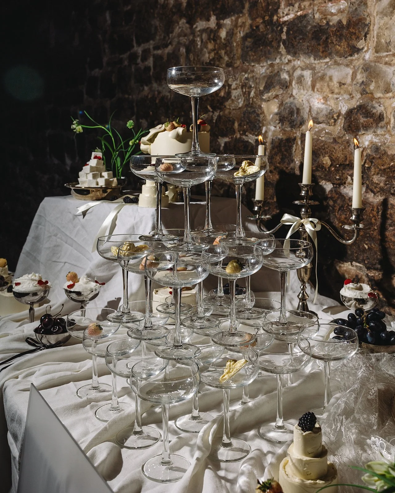 A pyramid of empty champagne glasses on a white tablecloth, with a stone wall and candles in the background, decorated for a celebration.