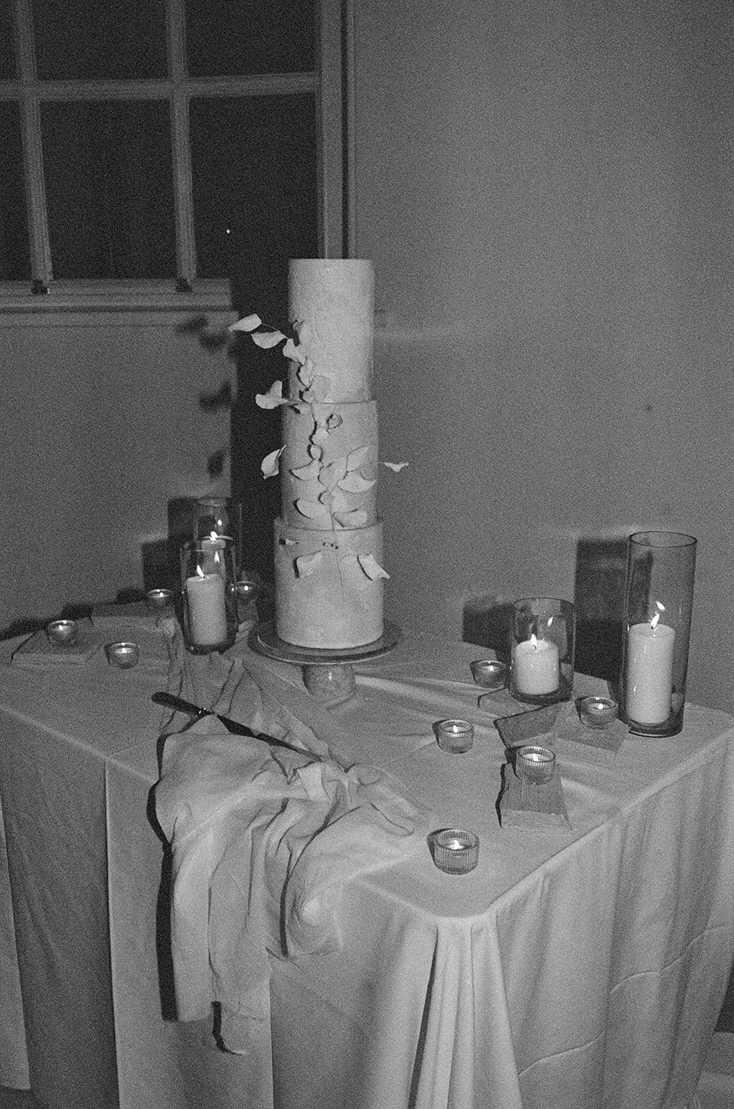 A table decorated with candles, a tall white cylindrical vase with paper flowers, and a draped cloth, in a dimly lit room with a window in the background.