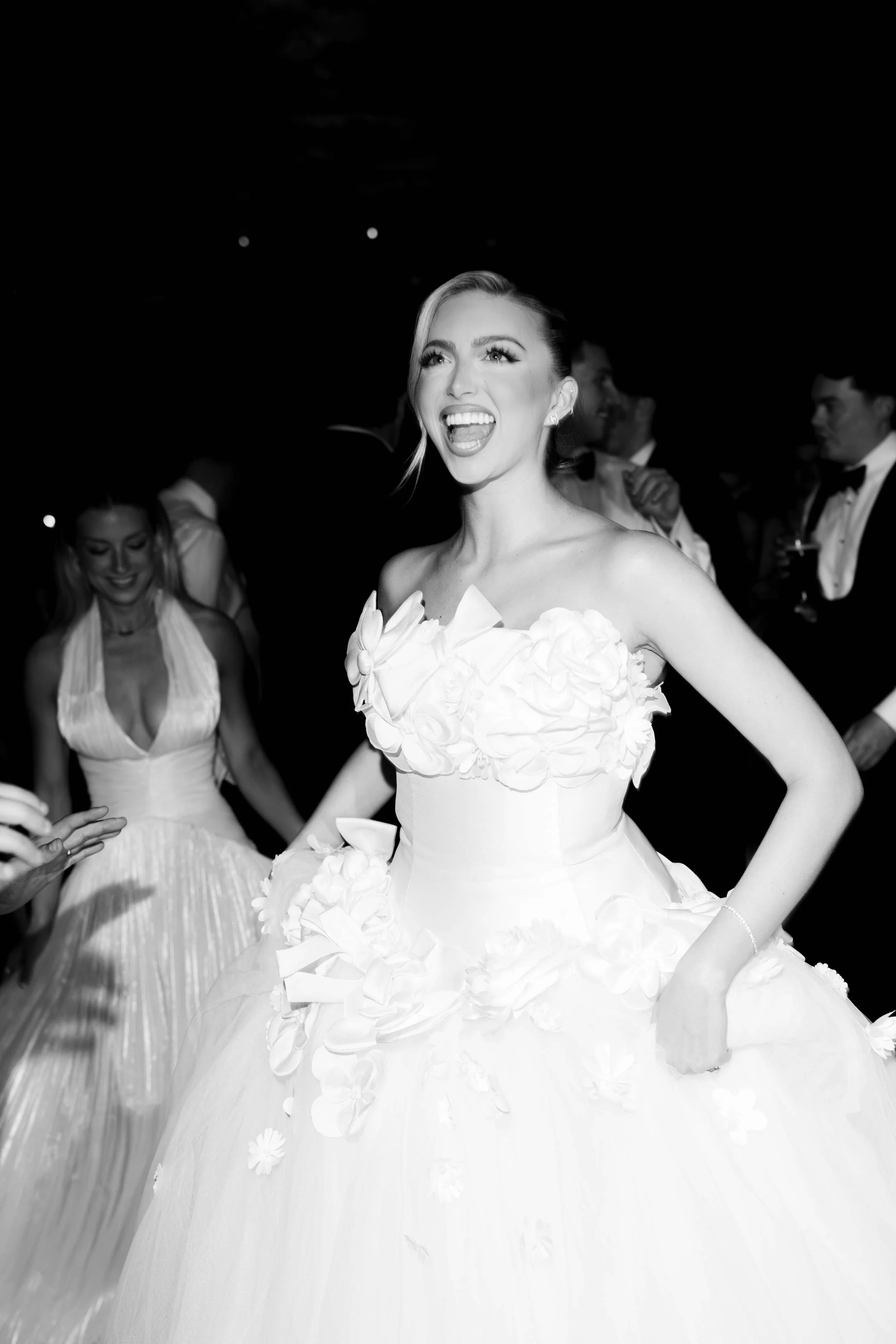 A woman in a wedding dress with floral details, smiling and sticking her tongue out, at a wedding reception with guests in the background.
