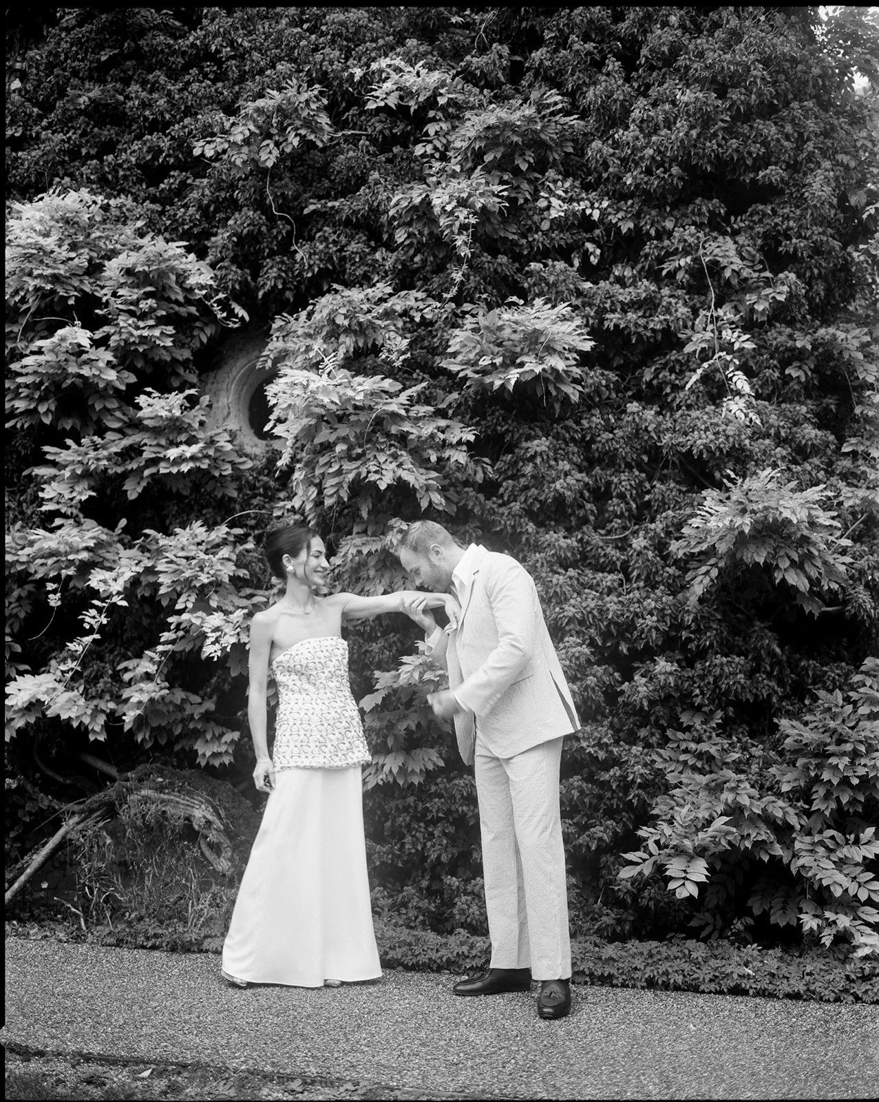 A couple in formal attire standing outdoors in front of lush greenery. The woman, wearing a strapless dress, is smiling while the man, in a light-colored suit, is leaning forward with his head bowed as he kisses her hand.