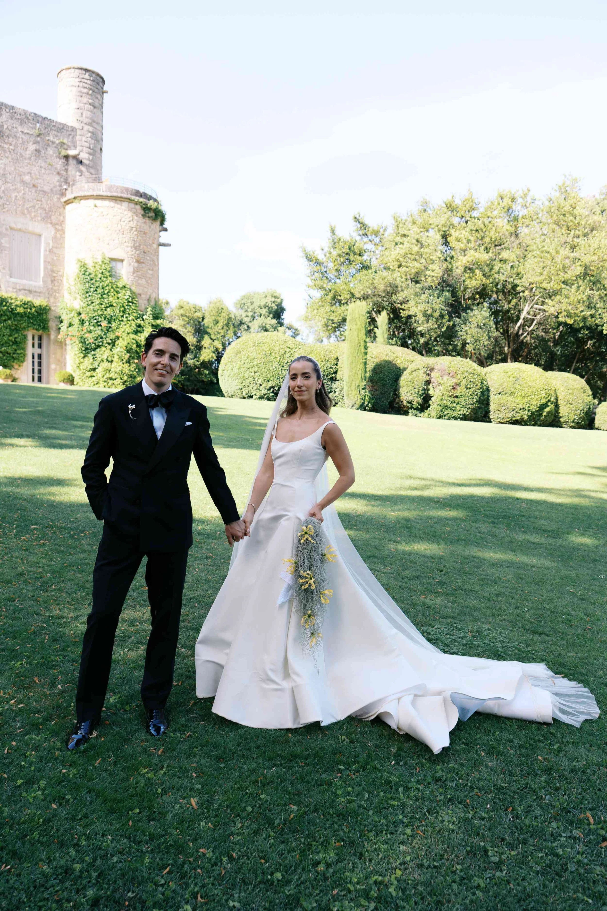 A bride and groom holding hands outside on a lawn with trimmed bushes and trees in the background, near an old stone building.