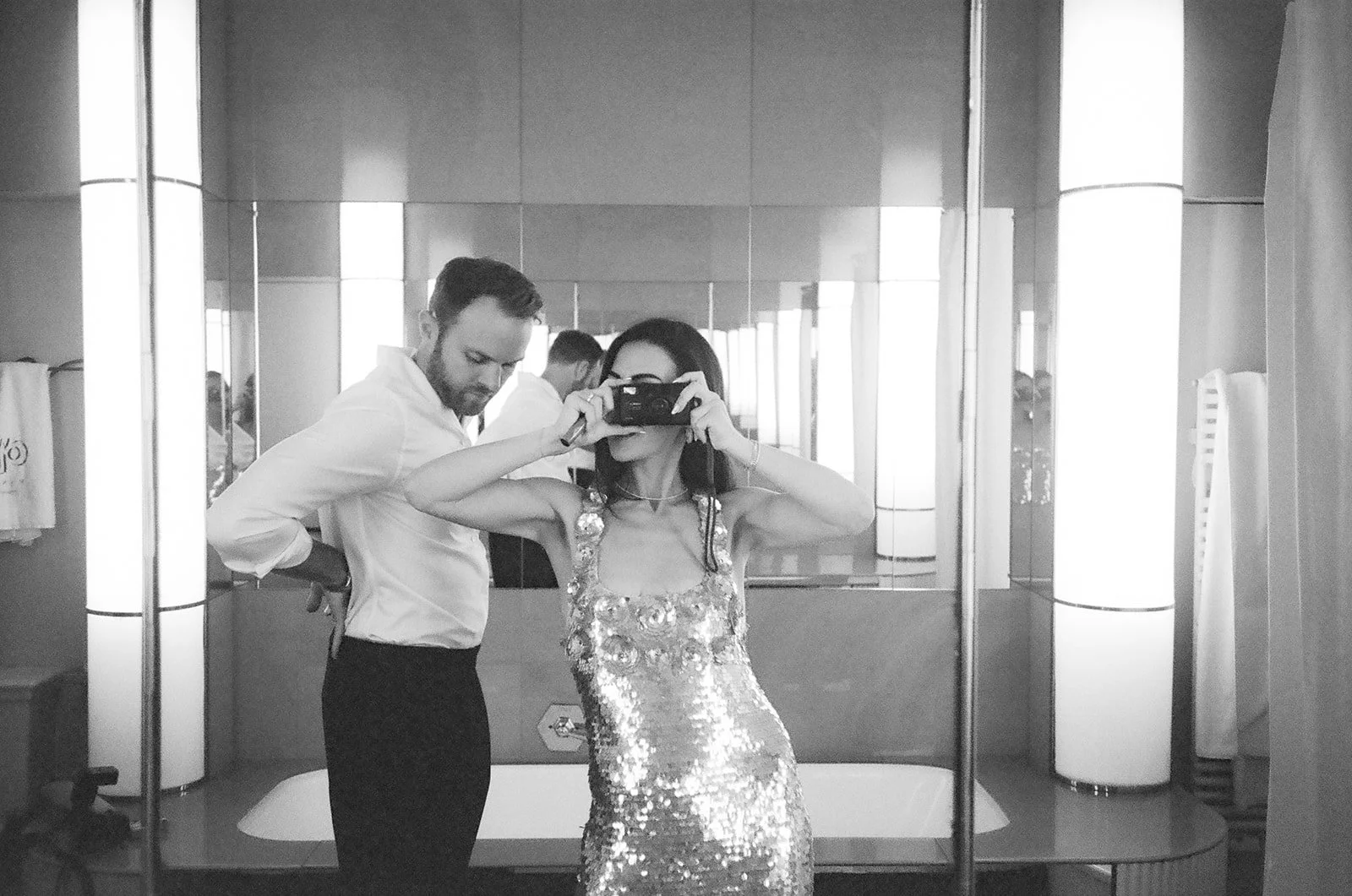 Black and white photo of a woman taking a selfie in a mirror, wearing a sequined dress, with two men, one with a beard, in a well-lit modern room with large mirrors and cylindrical light fixtures.