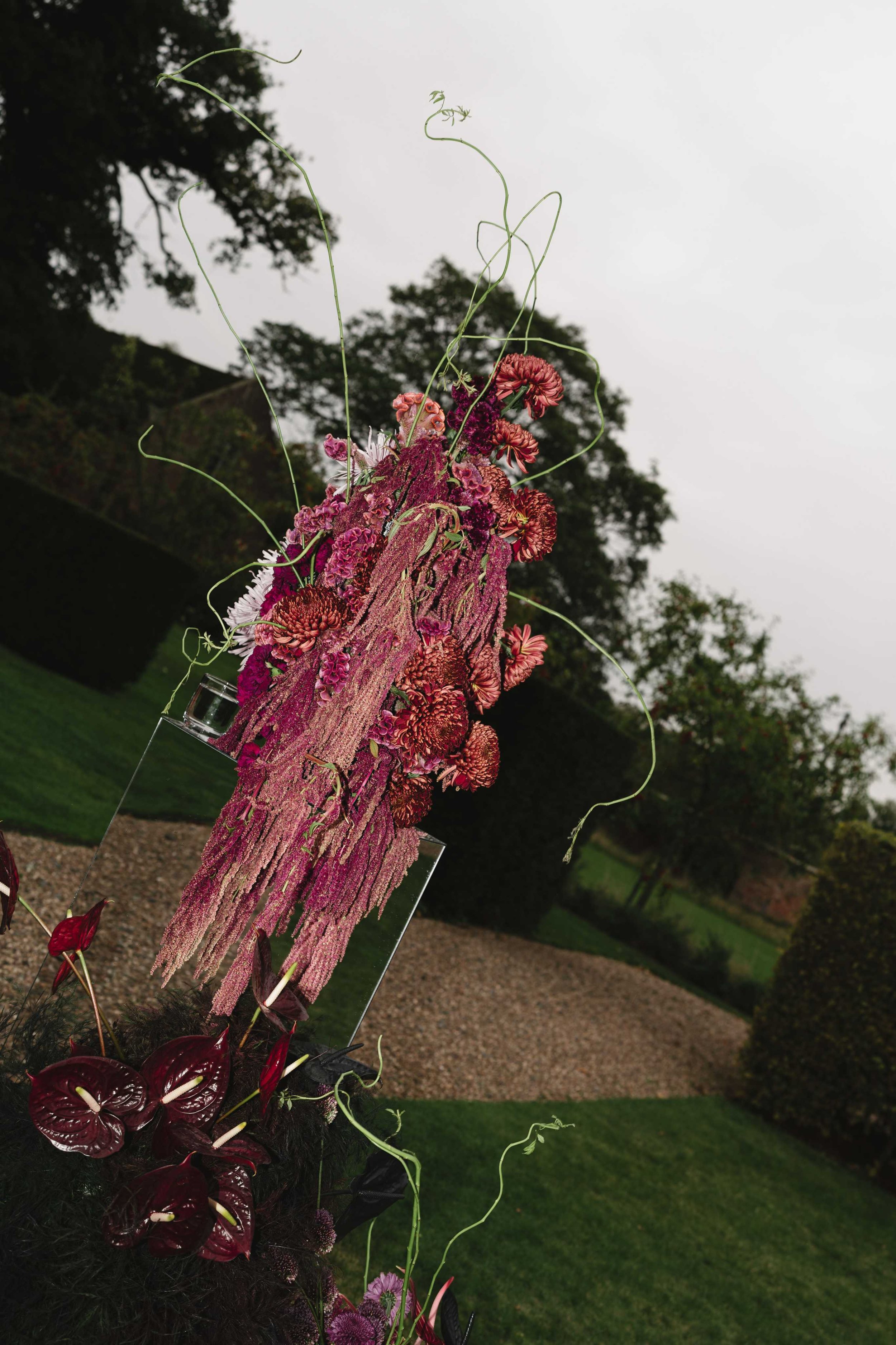A vertical arrangement of various flowers including pink, red, and purple blossoms, with long green tendrils and leaves, set against an overcast sky and a background of trees and grass.
