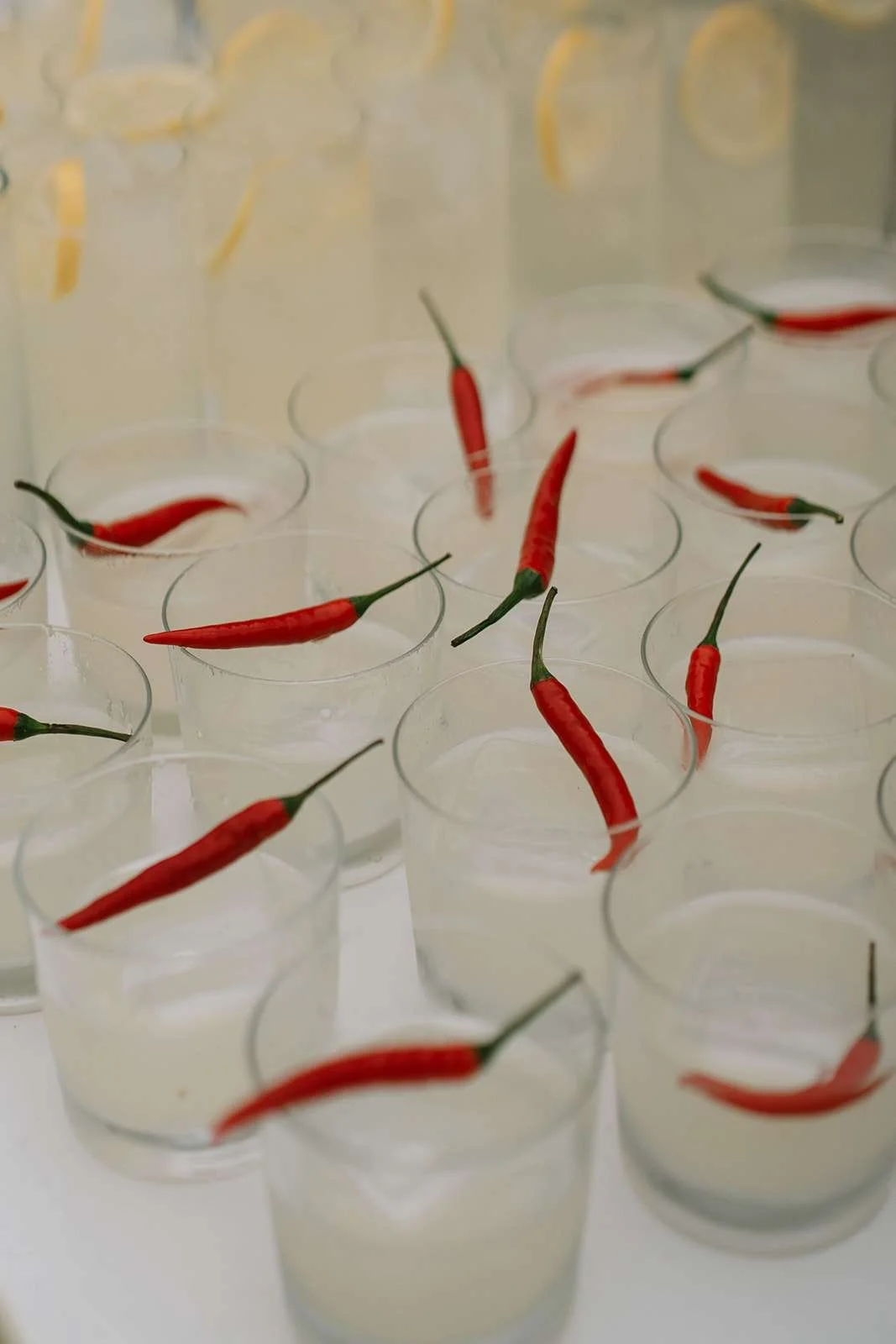 Multiple small clear plastic cups filled with a white liquid, each containing a red chili pepper, some with the chili submerged and others resting on the rim.