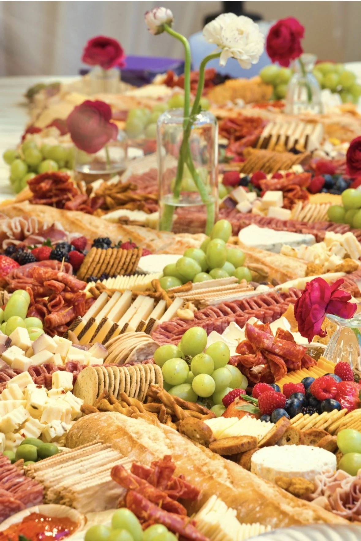 Grazing table with assorted fruits, cheeses, meats, crackers, and garnishes; view of a marina with boats and a skyline in the background.