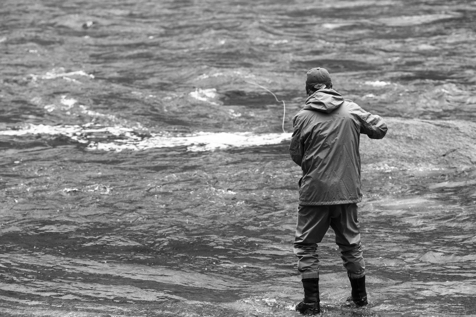 A person wearing waterproof clothing and boots standing in shallow water, fishing with a fishing line in hand, facing towards the water.