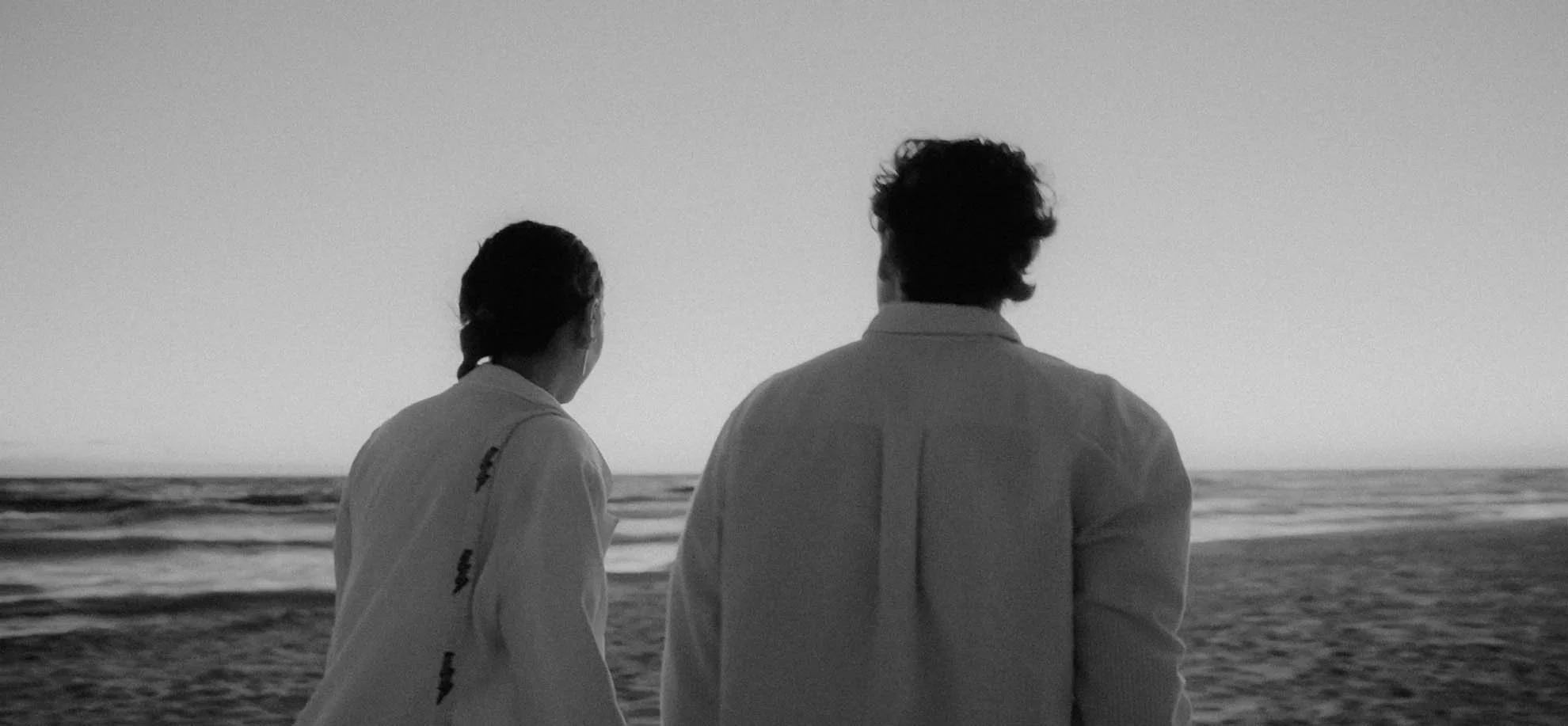 A black and white photo of a man and a woman standing on the beach, facing the ocean during sunset or sunrise.