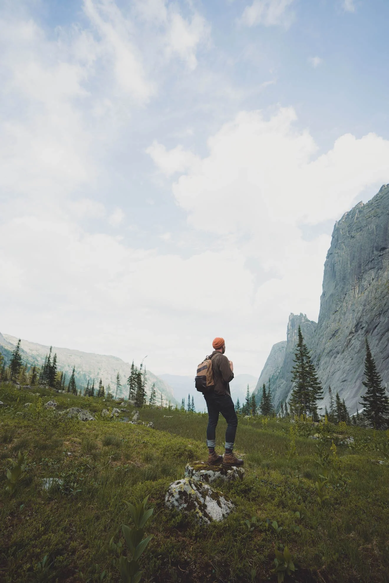 Hiker with backpack standing on a rocky foreground observing a mountain valley with tall cliffs and green trees under a partly cloudy sky.