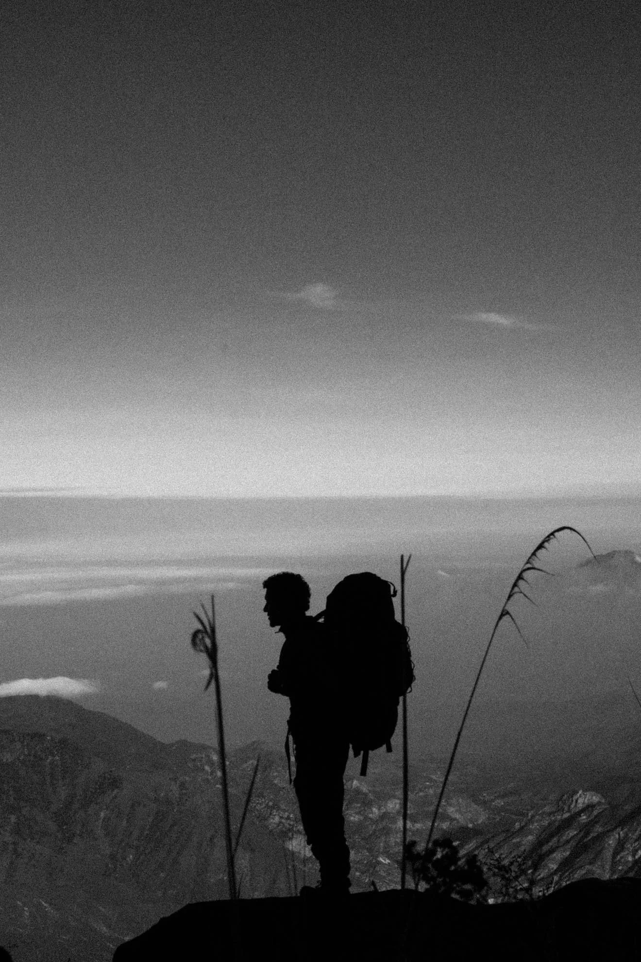 Silhouette of a hiker with a backpack holding hiking poles, standing on a mountain trail, overlooking a foggy valley and distant mountains with two small clouds in a black and white landscape.