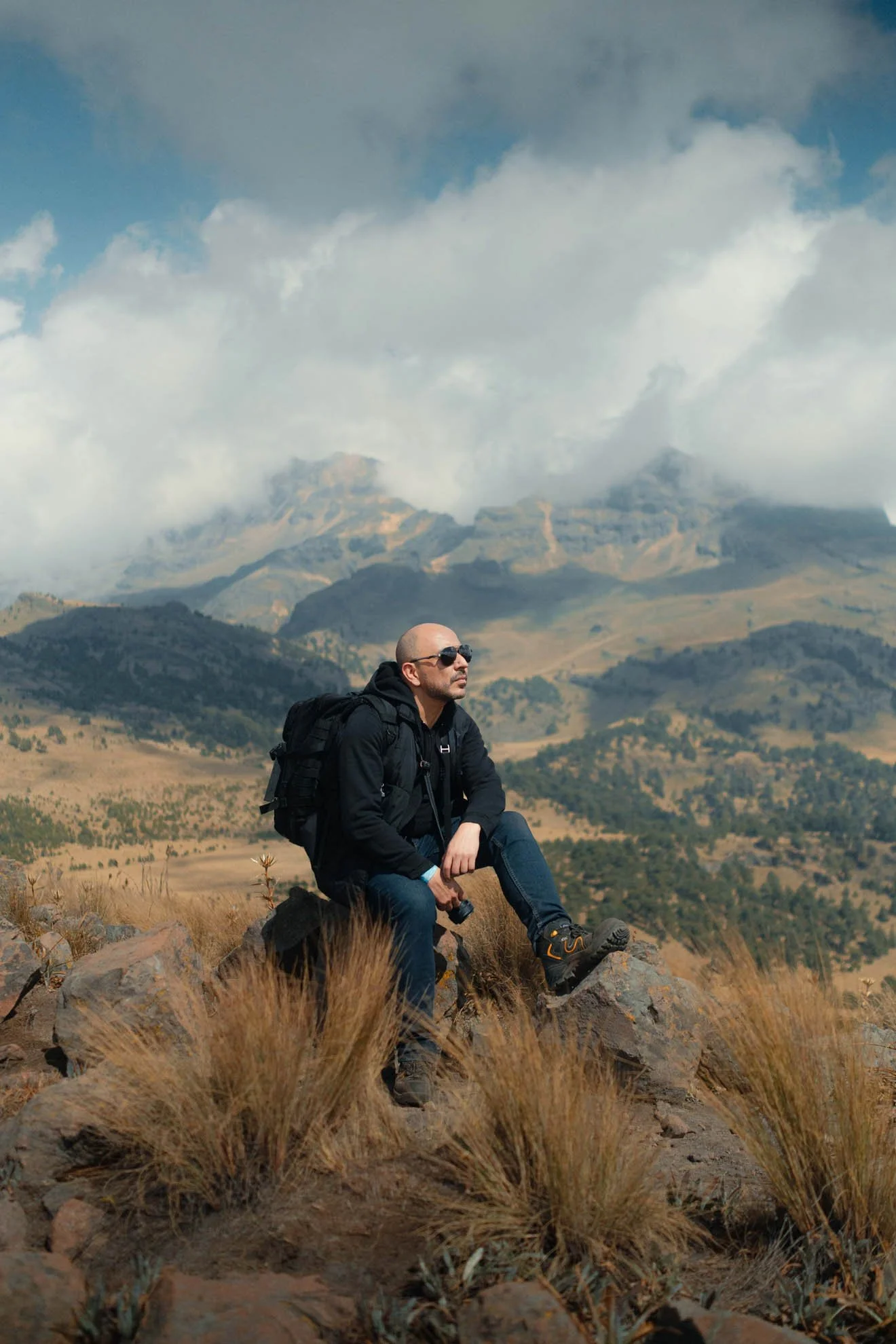 Man sitting on rocks in a mountainous landscape with cloudy sky, wearing sunglasses, black jacket, jeans, and hiking boots, holding a camera, with a backpack.