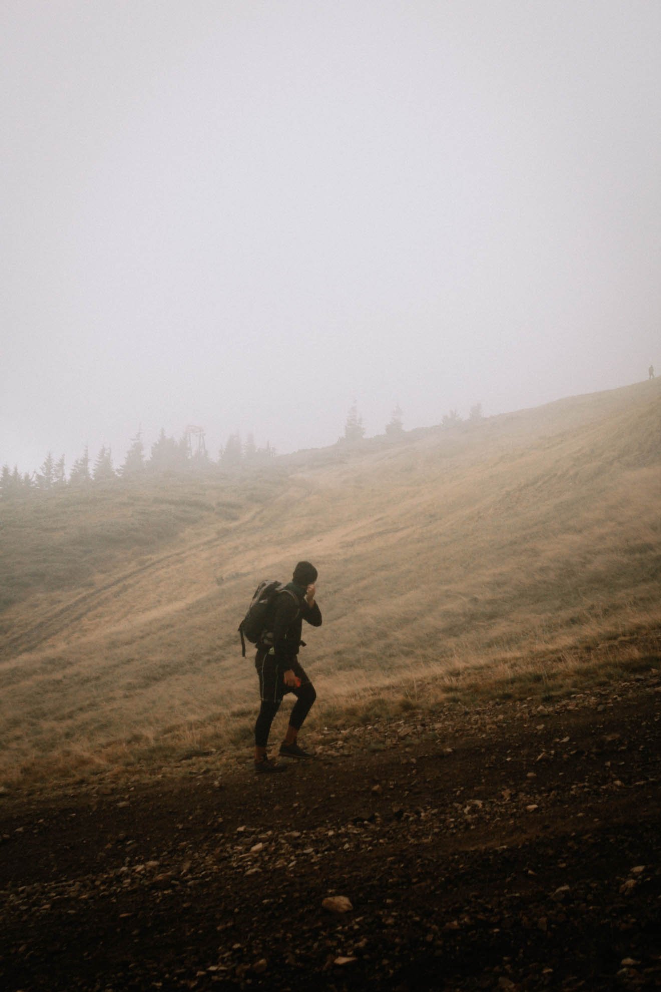 A person with a backpack hiking on a mountainous trail in foggy weather, with a lone figure visible in the distance against a misty sky.