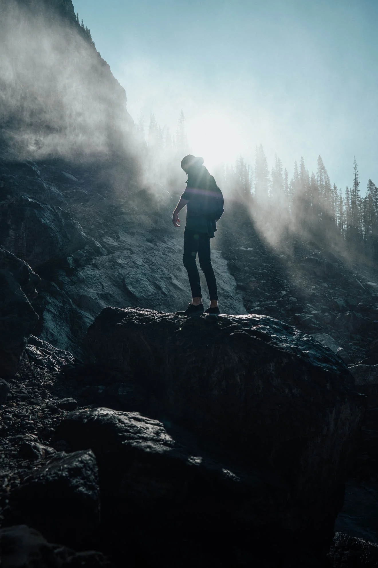 A hiker wearing a hat and backpack standing on rocks on a mountain trail with tall trees and misty mountains in the background, illuminated by sunlight.
