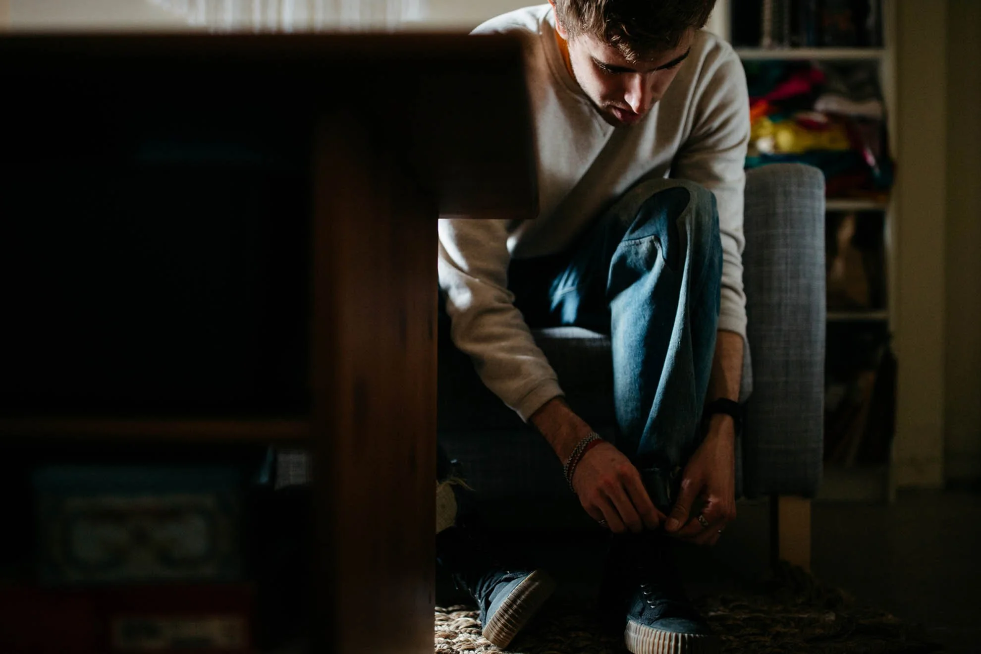 A young man sits on the floor next to a sofa, looking down with a focus on his feet, with a bookshelf in the background.